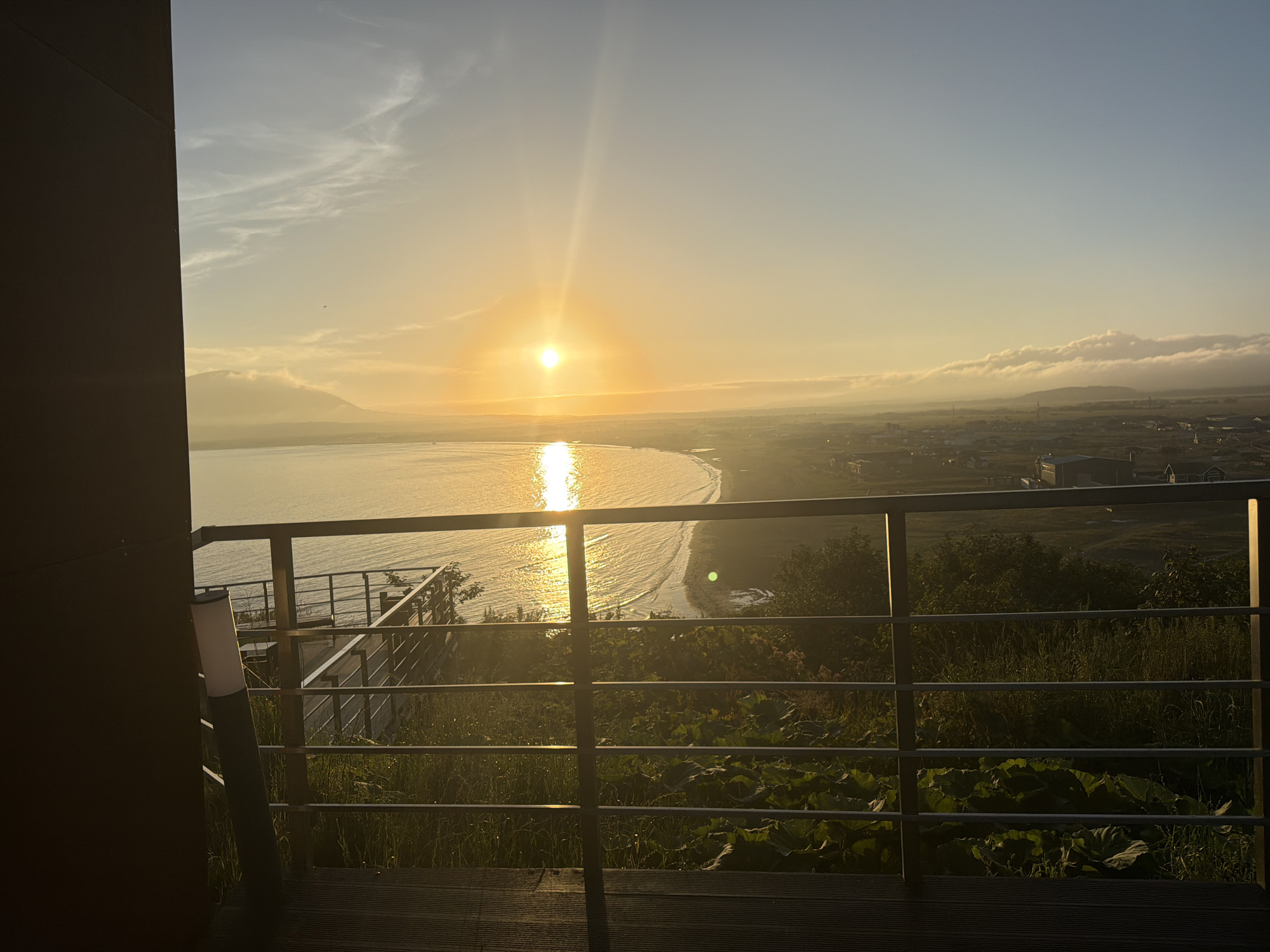 This image captures a sunrise over the Sea of Okhotsk from a vantage point in Yuzhno-Kurilsk, Russia. The sun, a bright orb, is positioned low on the horizon, casting a golden glow across the water and sky. Its reflection creates a shimmering path on the gently rippling sea. A curved coastline is visible, bordered by a strip of land with scattered buildings and vegetation. In the foreground, a metal railing and lush green plants suggest a balcony or observation deck. The atmosphere is serene, with soft clouds streaking across the pale blue sky, and a distant mountain range is faintly discernible through the morning mist.