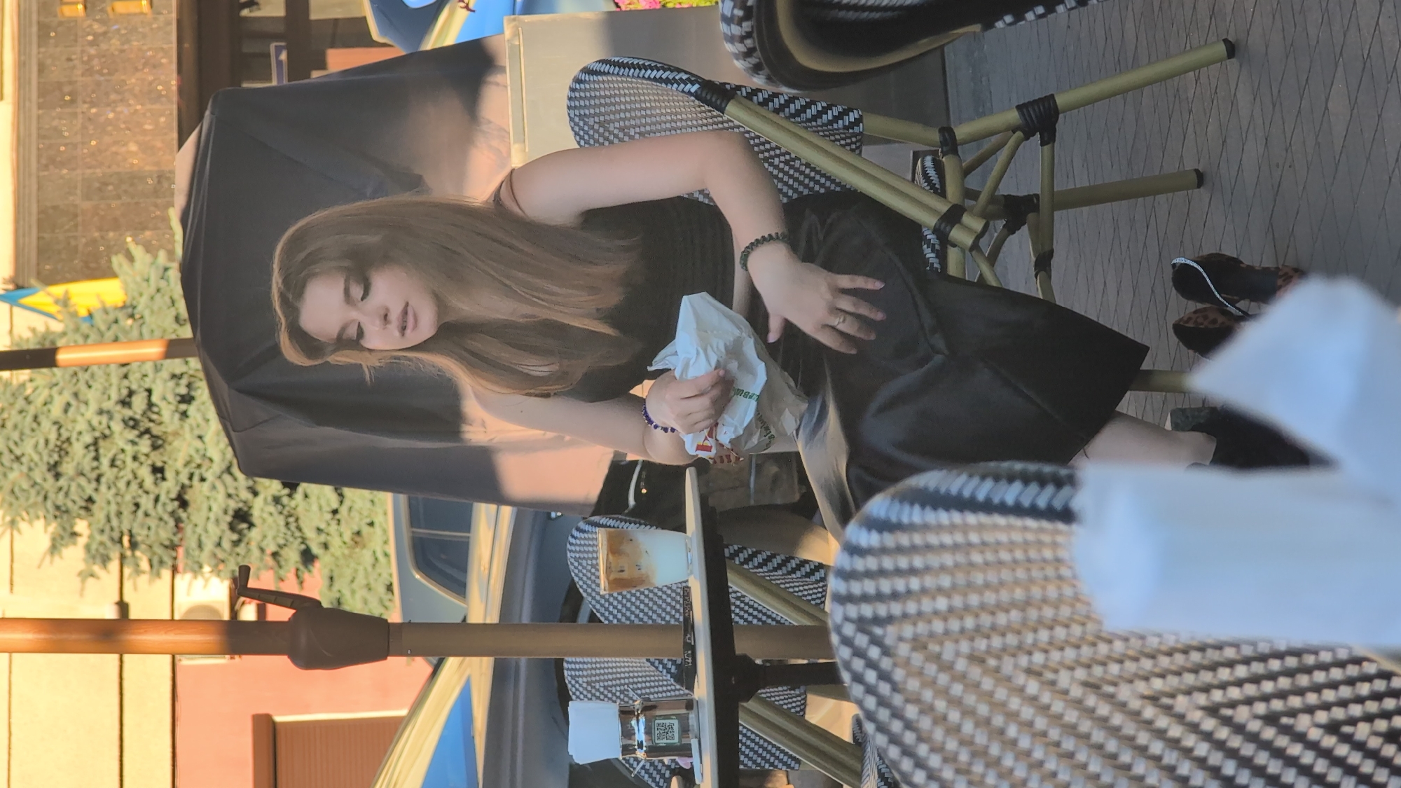 A young woman with long, light brown hair is seated at an outdoor cafe table. She is wearing a black top and a black skirt. Her eyes are closed, and she appears to be resting or perhaps sleeping. She holds a white paper bag in her hands, which she is clutching. On the table in front of her is a glass with a light-colored beverage, possibly coffee with cream. There is also a small bottle with a green label on the table. The cafe setting is indicated by the chairs with a black and white woven pattern and the presence of an umbrella for shade. In the background, there are trees and buildings. The lighting suggests it is daytime, likely late afternoon due to the warm, golden hue of the sunlight. The woman has jewelry on her wrist, including bracelets and a ring on her finger. The overall scene conveys a sense of relaxation and casual enjoyment of a cafe experience. There are no discernible text elements in the provided image, other than a faint indication of a brand name on the paper bag which is illegible.