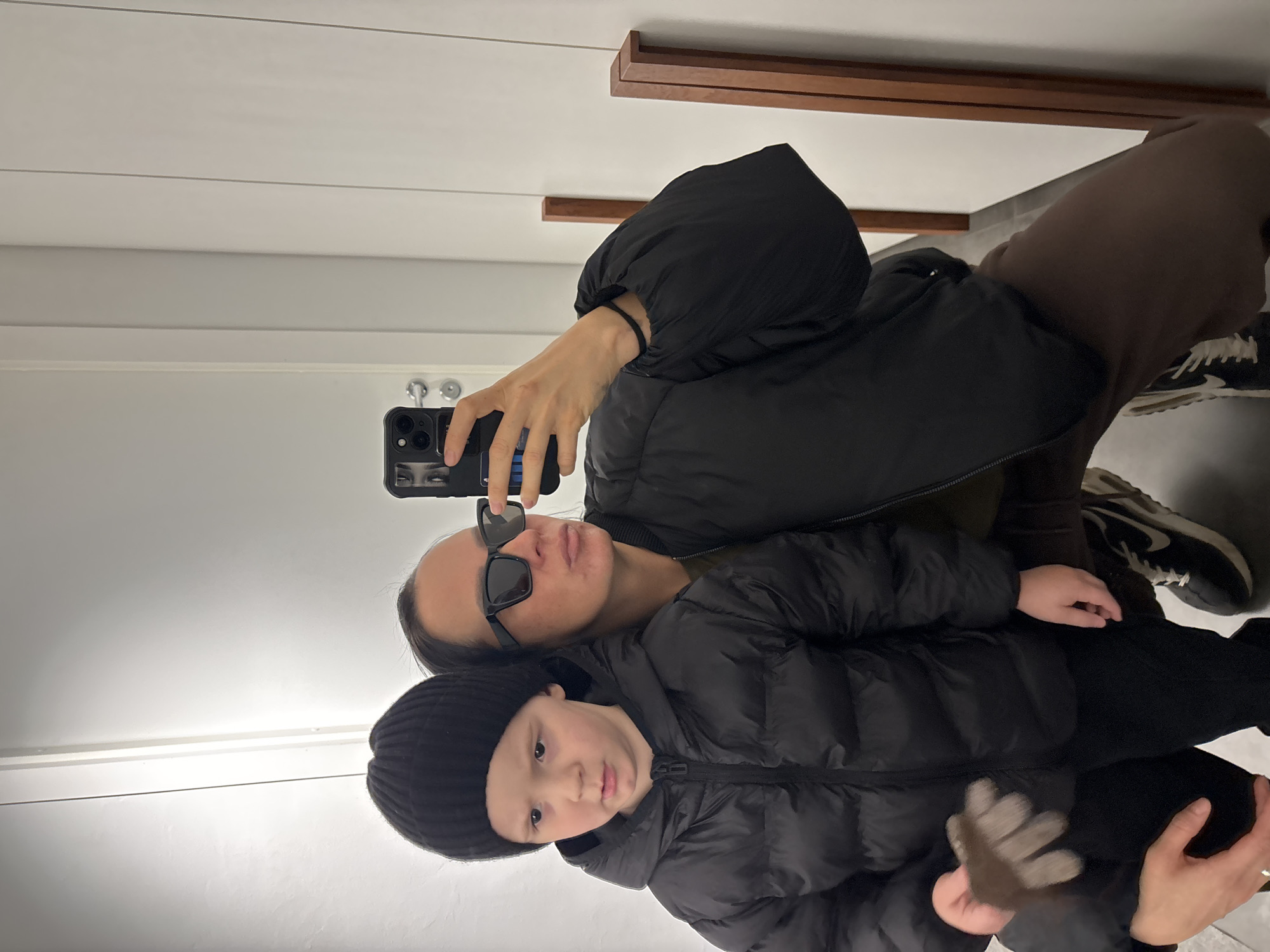 A selfie photograph shows a woman and a young child in a mirror. The woman is wearing sunglasses and a black puffer jacket with a hood. The child is wearing a black beanie and a black puffer jacket. The woman is holding a smartphone and taking the photograph. The location is likely a bathroom, as evidenced by the mirror and the white walls. The city of Chaikovsky, Russia, is not discernible from the image.