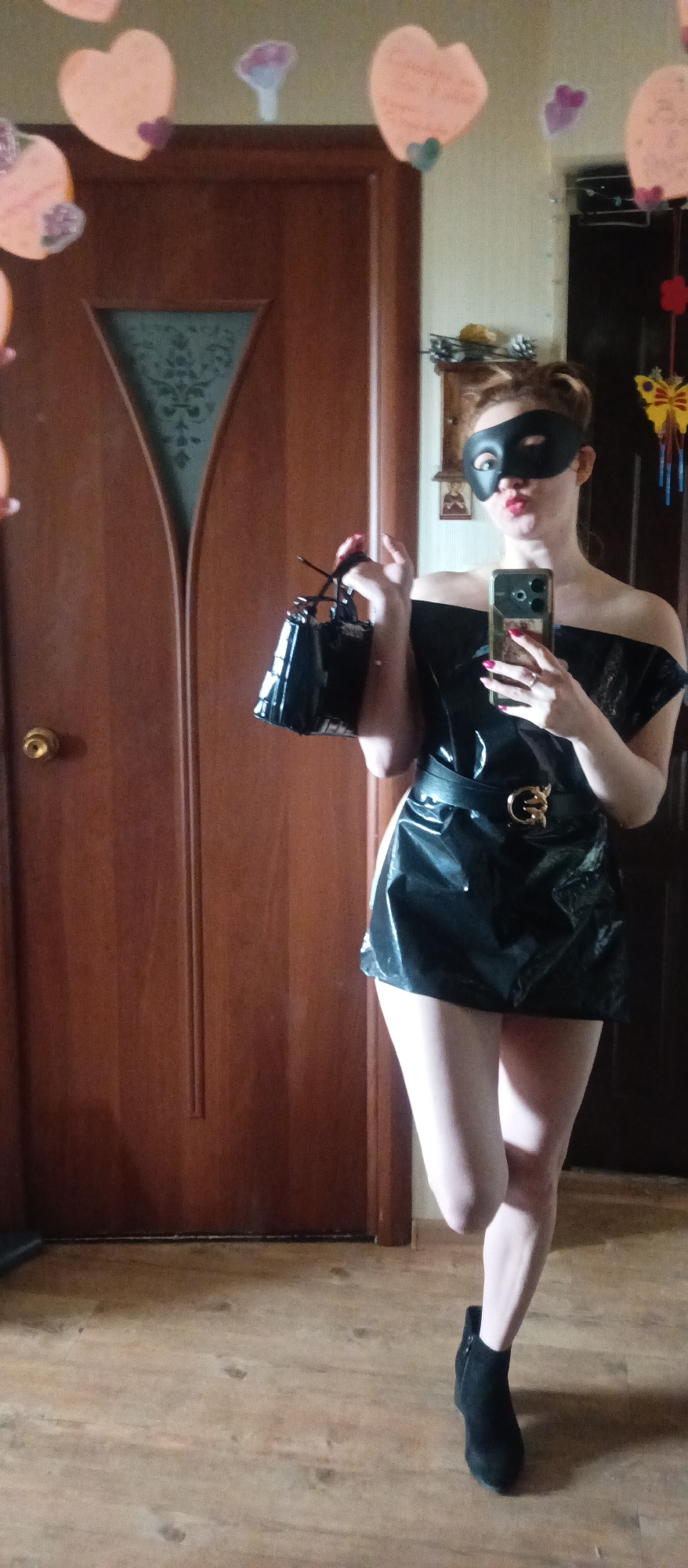 A full indoor shot depicts a fair-skinned woman in a black mask. She holds a shiny black purse in her right hand and a cell phone in her left. Her hair is piled up. She wears a shiny black off-the-shoulder dress with a black belt and buckle. She wears skin-tone tights and black ankle boots. She stands in front of a wooden door in a room. The floor is wooden. The wall has colorful decorations. There is a mirror on the wall with heart shapes attached. Location: Unknown, Unknown.
