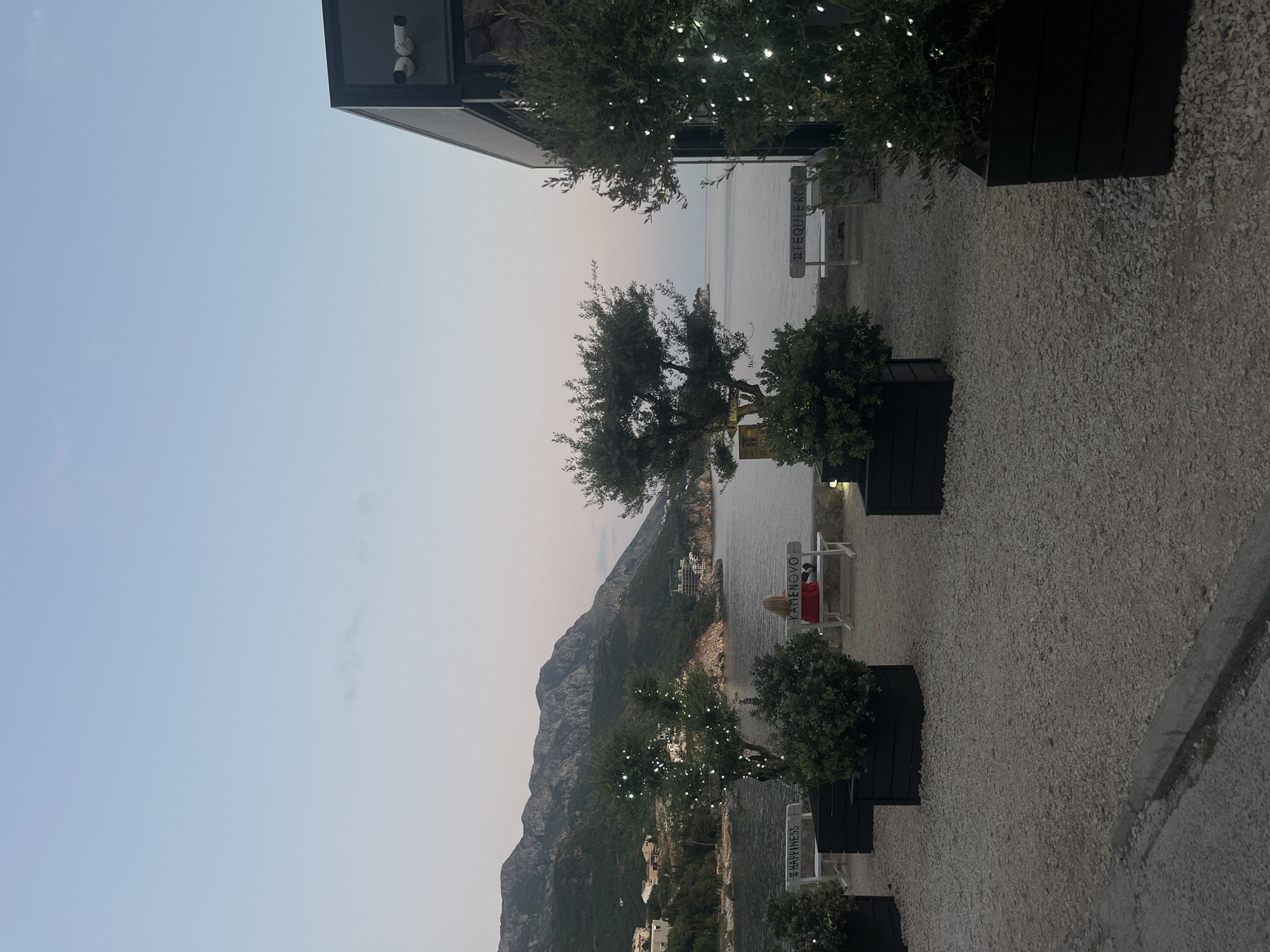 The image depicts a coastal scene with a view of mountains and the sea. A person is sitting on a bench with the words "KAMENOVO" written on it. There are potted trees decorated with lights along a pathway covered with gravel. In the background, a mountain range is visible. The sky is clear and light-colored. The location is in the city of Unknown, Unknown.