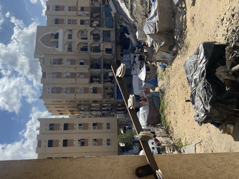 This image captures a devastated urban landscape in Khan Yunis, Palestinian Territory, under a partially cloudy blue sky. The scene is viewed from an elevated position, with a portion of a beige building wall and a horizontal metal bar featuring two white ceramic pulleys visible on the immediate left foreground.
The midground consists of a large, sandy, and debris-strewn open area. This space is occupied by numerous makeshift shelters of varying sizes and constructions, fashioned from tarpaulins, fabric sheets, and other salvaged materials. Patches of sparse green vegetation are interspersed among the sand and rubble. In the lower right, a substantial pile of debris is covered by a black tarpaulin.
In the background, multiple multi-story buildings exhibit severe structural damage. The central and rightmost buildings, constructed of light brown material with darker accent bands and an arched decorative element on the central building's upper facade, show extensive destruction. Lower and middle floors are significantly compromised, with large sections of walls missing, exposing interior structures and piles of rubble. The facades of all visible buildings are heavily marked with numerous small impact holes, and many windows are broken or missing. Some windows on less damaged upper floors display items hanging from them. The overall environment indicates widespread destruction and the presence of temporary settlements. No individuals are visible in the frame.