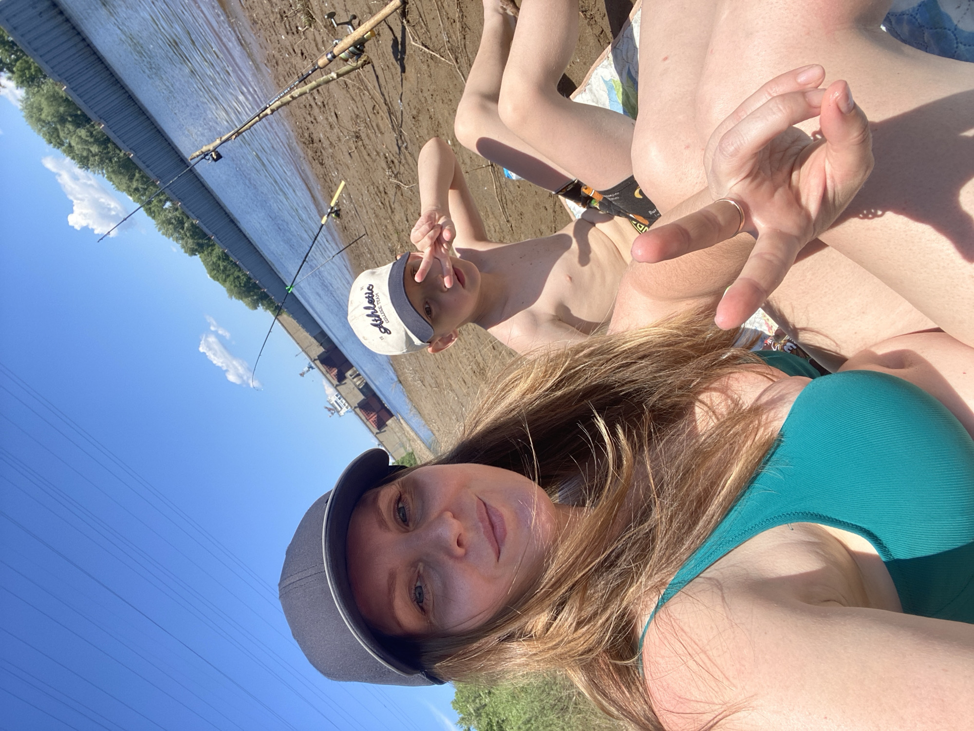 This is a close-up, selfie-style photograph taken outdoors on a sunny day, likely on the sandy bank of the Kama River near Chaikovsky, Russia. In the foreground, a woman wearing a dark baseball cap and a teal bikini top is partially visible, smiling at the camera. To her right, a young boy wearing a light-colored baseball cap with the word "Athletic" visible, and patterned swim trunks, is also in the frame. He is making a peace sign with his right hand. The background shows the wide expanse of the Kama River, with trees lining the opposite bank. Several fishing rods are set up on the riverbank, indicating fishing activity. A large industrial structure, possibly a dam or part of a port, is visible in the distance across the river. The ground is sandy and somewhat uneven, with some sparse vegetation. The sky is a clear blue with a few scattered white clouds.