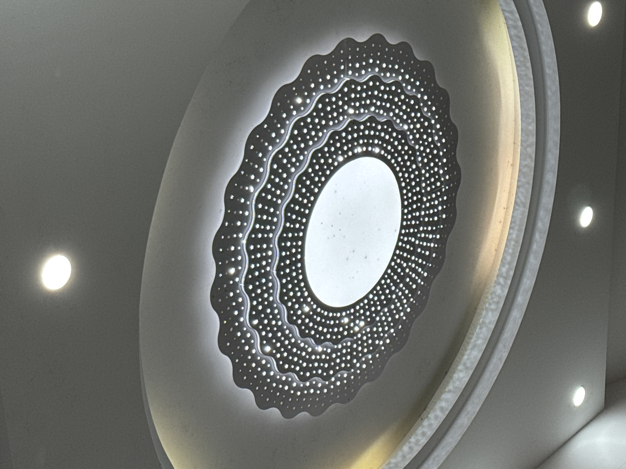 The image displays an upward-angled view of a decorative ceiling light fixture in an indoor setting within Wulingyuan, China. The central subject is a large, oval-shaped fixture recessed into a multi-tiered ceiling. This fixture features a bright, translucent oval panel at its center, surrounded by several concentric, dark-colored rings with a wavy, scalloped edge. These rings are intricately perforated with numerous small, bright, star-shaped and circular LED lights, creating a sparkling effect. The fixture is illuminated. Surrounding the main fixture are additional, smaller, circular recessed spotlights embedded in the white or light-grey ceiling, three of which are visible and illuminated, providing ambient light. The overall scene shows a modern, well-lit interior ceiling. No individuals or specific actions are depicted.