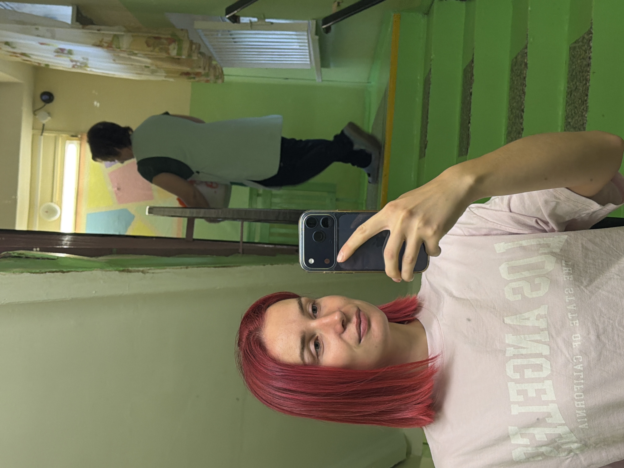 In the city of Chaikovsky, Russia, a young woman with vibrant pink hair is taking a selfie in a hallway. She is wearing a light pink t-shirt with "LOS ANGELES" and "THE STATE OF CALIFORNIA" printed on it. In the background, an older woman wearing a light green vest over a dark green shirt and black pants walks down a bright green staircase, carrying a white bag. The hallway features green painted walls and stairs, a window with a colorful decoration, and a white metal railing.