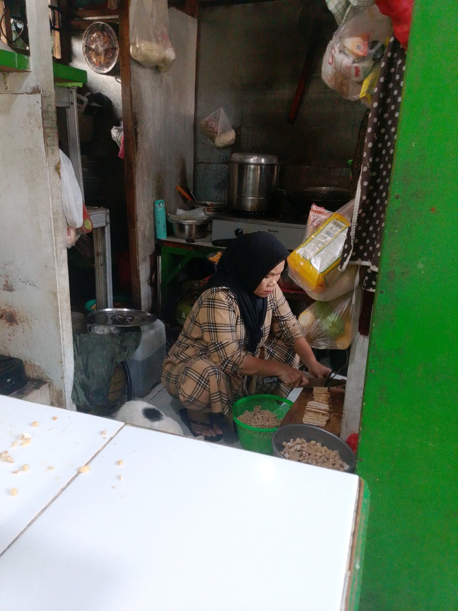 The image depicts a street food vendor preparing food in a small, cluttered outdoor stall in East Jakarta, Indonesia. A woman, wearing a black hijab and a plaid patterned outfit, is crouched down and actively slicing pieces of food on a wooden cutting board. She is using a knife in her right hand to cut what appears to be tempeh. In front of her, to her left, is a green plastic basket filled with what looks like chopped tempeh, and to her right, a grey bowl containing more chopped tempeh. Behind her, various kitchen items, including stainless steel pots and plastic bags of ingredients, are visible. The stall itself is simple, with concrete walls and a corrugated metal roof. The foreground shows white surfaces, possibly tables or counters, with some food scattered on them. A small white and black dog is partially visible in the lower center of the image, near the woman's feet. The overall environment suggests a busy, functional food preparation area within a densely populated urban setting.