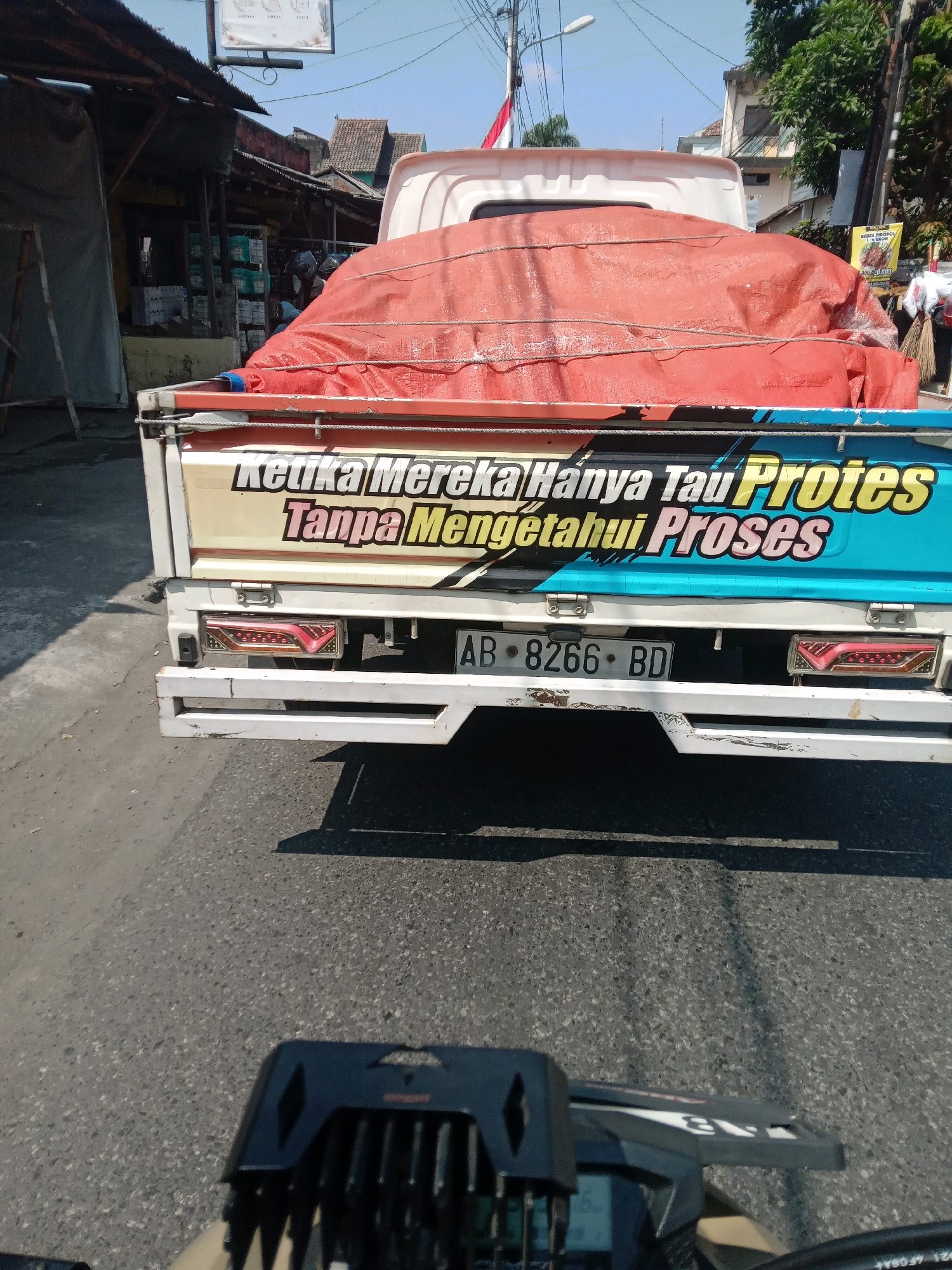 The image shows the rear of a white truck with a red tarp covering its load. The truck has colorful lettering on its side that reads "Ketika Mereka Hanya Tau Protes Tanpa Mengetahui Proses" in yellow and blue. A license plate reading "AB 8266 BD" is visible. The truck is on a paved road, likely in the city of Condongcatur, Indonesia, based on the style of the buildings and the overall environment. The scene appears to be during the daytime, with clear skies. In the foreground, the handlebars and part of the dashboard of a motorcycle are visible, suggesting the photo was taken from a motorcycle. There are buildings and shops on the left side of the road, and trees and other buildings in the background. The lighting indicates a sunny day.