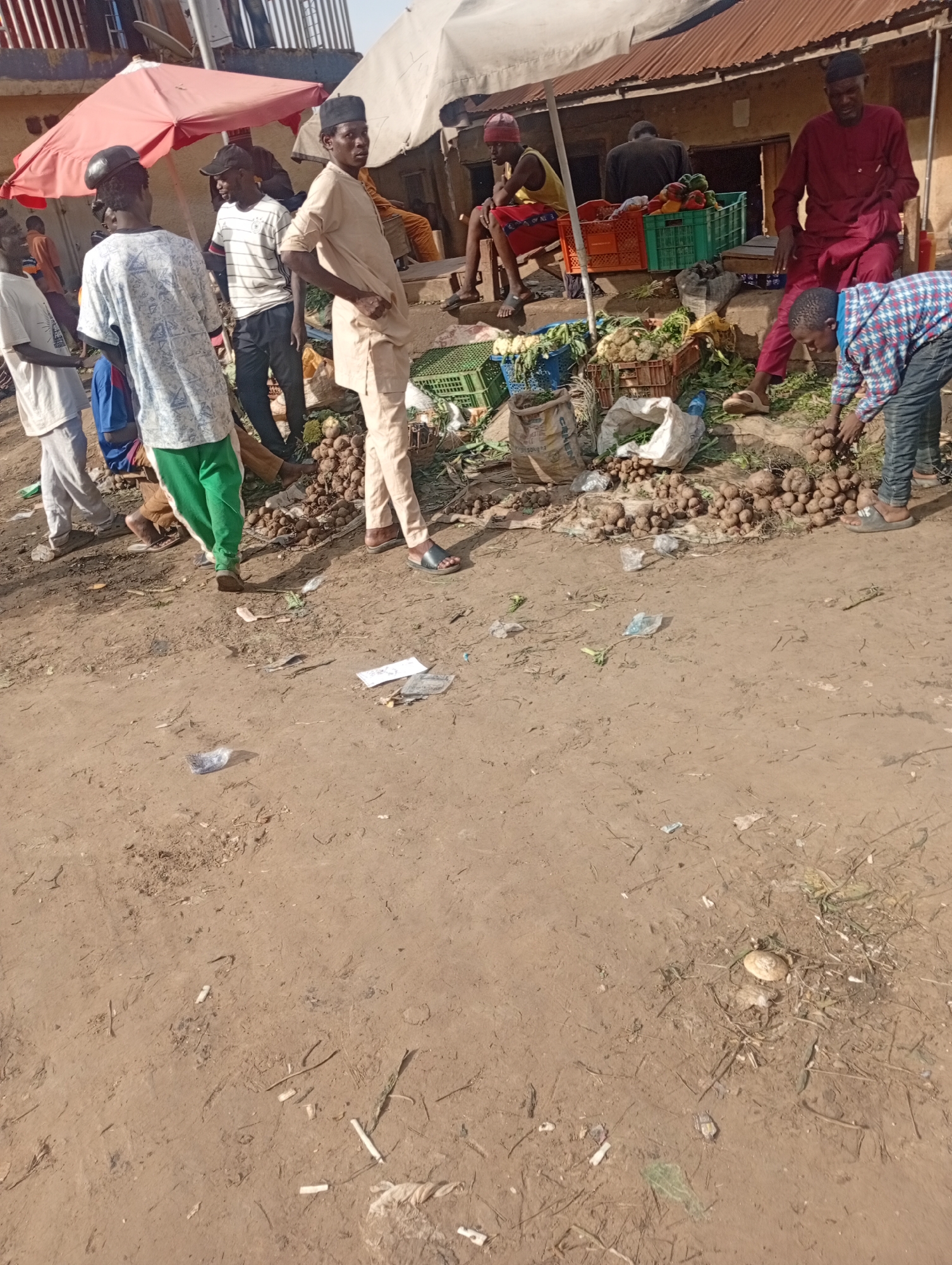 The image depicts an outdoor market scene in Rafin Par, Nigeria. The ground is dusty and unpaved, with scattered debris including plastic wrappers and dried vegetation. In the background, there are stalls with corrugated metal roofs. Several individuals are present, engaged in what appears to be trade or transaction.
On the left side of the image, a person in a light-colored shirt and green pants is walking. Near them, another person in a patterned shirt and light-colored pants stands with their hands on their hips. A man wearing a light brown tunic and a black cap stands in the center, facing the camera. To his right, another man in a burgundy outfit and a black cap is seated. In the foreground, a person in a checkered shirt and blue jeans is leaning down, handling what appear to be yams or similar root vegetables.
Various goods are displayed, including crates of produce like bell peppers, and large piles of root vegetables on the ground. Bags of produce are also visible. The overall atmosphere suggests a busy marketplace environment.