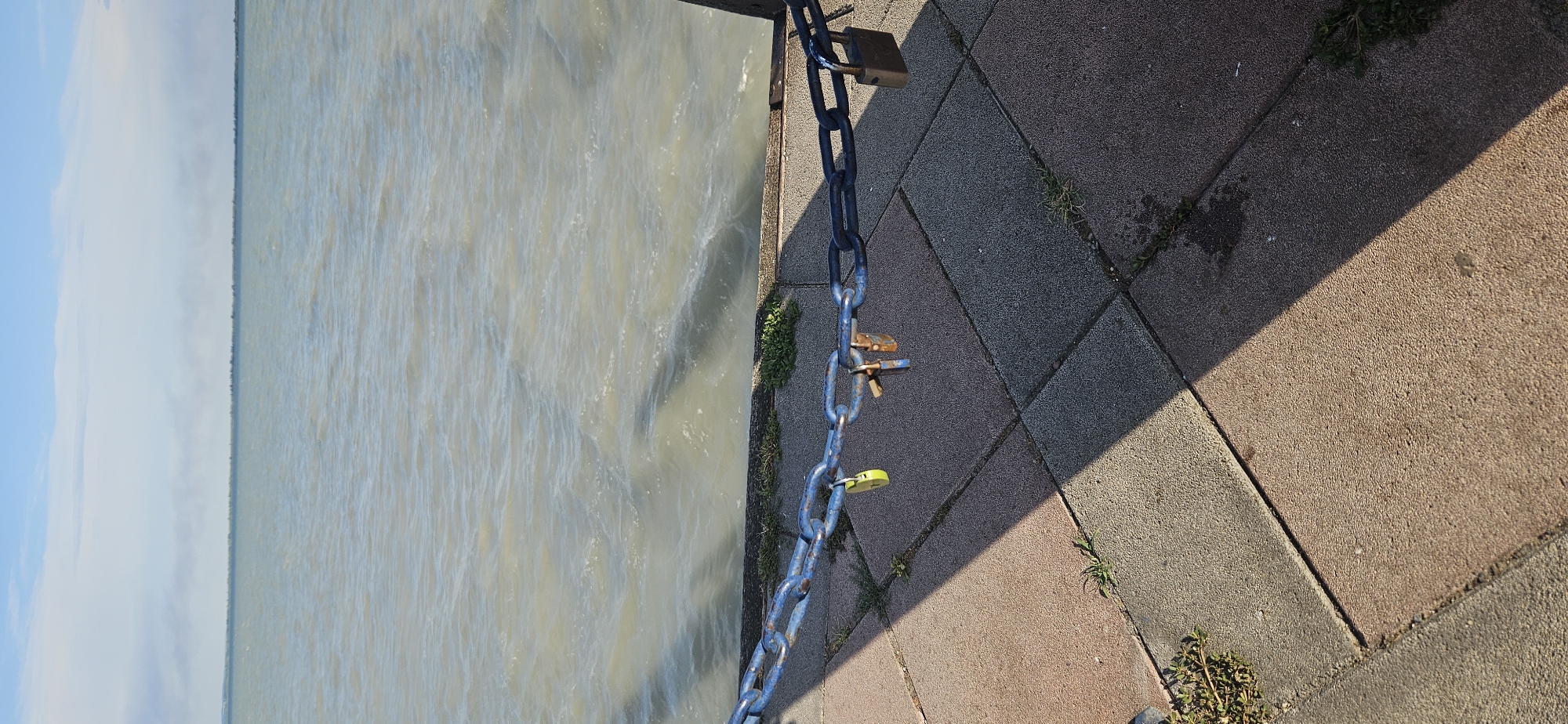 A chain secured with multiple padlocks stretches horizontally across the foreground of the image. The chain is attached to a wooden post on the right and extends out of frame to the left. The chain is linked to a pier or walkway, made of interlocking pavers in shades of pink, gray, and tan. Small patches of green grass grow between the pavers. In the background, a large body of water, likely Lake Balaton, stretches to the horizon under a pale blue sky with wispy clouds. The water is choppy, with visible ripples and waves. A thin line of trees or vegetation is visible on the distant shore. The scene is captured from a low angle, looking out over the water. The overall impression is one of a serene, outdoor location, possibly a public access point to the lake.
