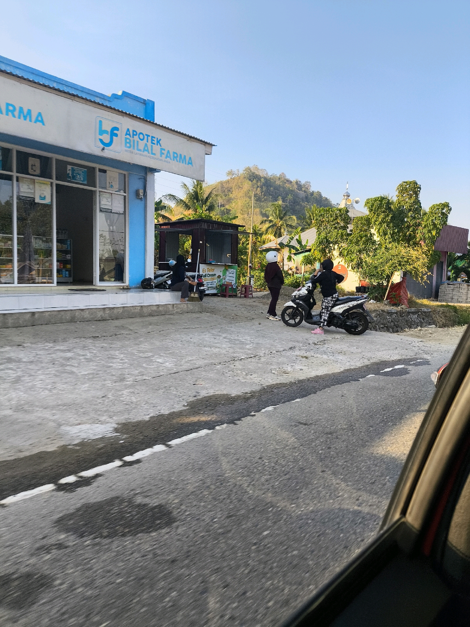 The image depicts a street scene in Dulupi, Indonesia, in front of a pharmacy called "APOTEK BILAL FARMA". The pharmacy has a blue and white facade with large glass windows showcasing products inside. Two individuals are present outside the pharmacy. One person, wearing a white helmet and dark clothing, stands on the sidewalk. Another person, on a motorcycle, is wearing a black top, patterned pants, and pink shoes. The motorcycle is white and black. In the background, a mosque with a silver dome and speakers is visible, surrounded by lush green trees and a hillside. The street in the foreground is paved with asphalt and shows signs of water or oil stains. A portion of a car interior, likely the window frame, is visible in the lower right corner, suggesting the photo was taken from inside a vehicle. The sky is clear and blue.
