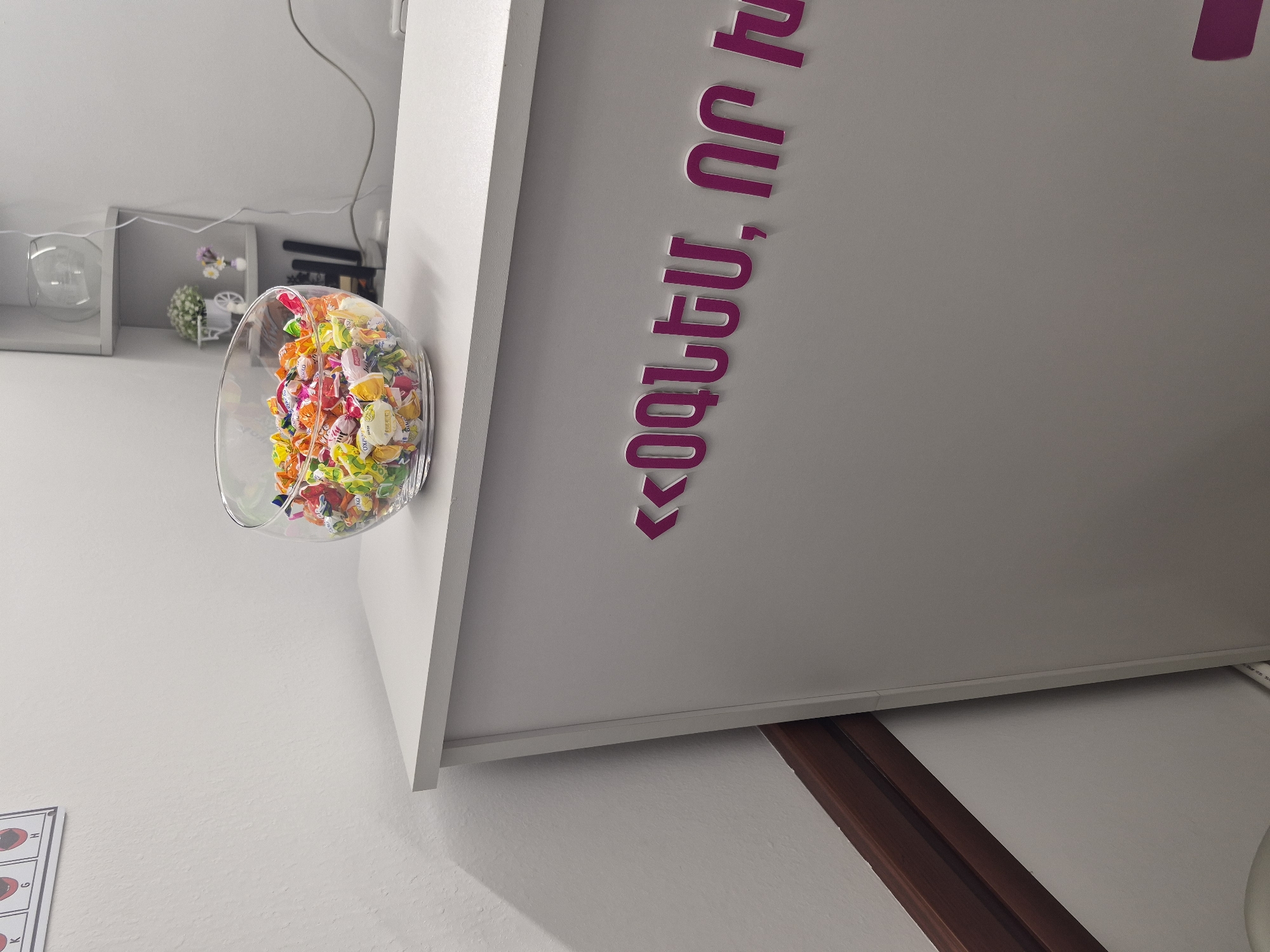 A close-up shot captures a white counter with a glass bowl filled with colorful, individually wrapped candies. The candies are a mix of red, orange, yellow, green, and white, and some wrappers are partially visible with branding. Behind the counter, on the wall, are large, raised magenta letters in Armenian script, with the first part appearing as "<<ՕԳՆԵՄ, ՈՐ Ի" which roughly translates to "I will help, that I". To the left of the counter, a sliver of a poster with phonetic symbols is visible on a white wall. The environment appears to be an indoor reception area or a waiting room. Given the Armenian script, this scene is likely located in Yerevan, Armenia.
