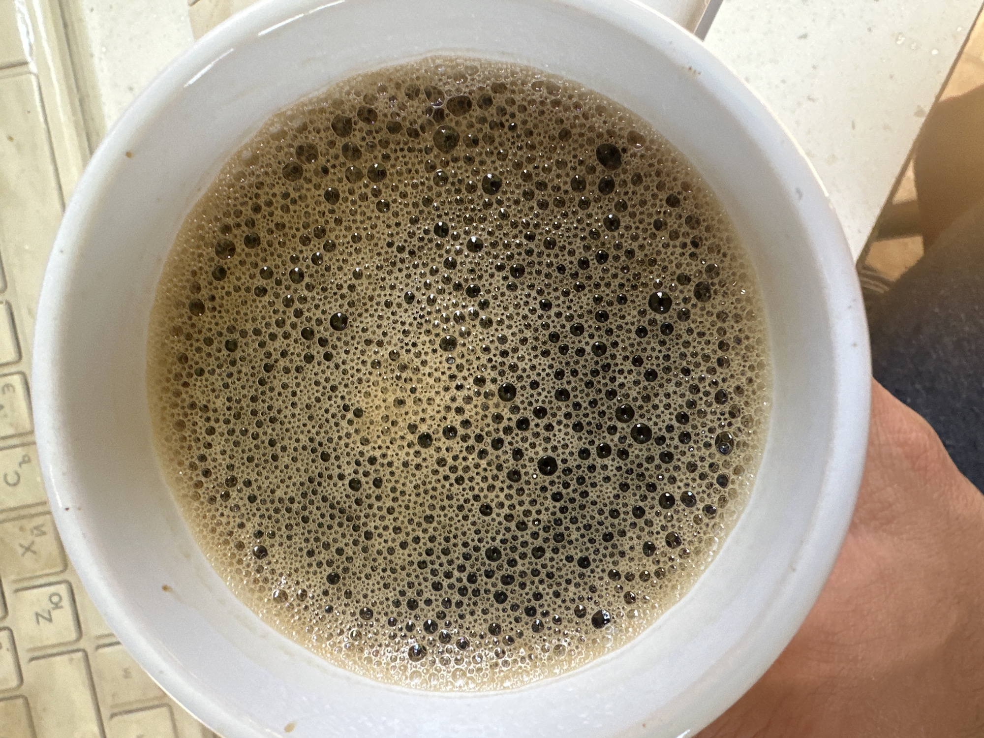 A top-down shot reveals a white ceramic mug filled with coffee, notably topped with a dense layer of dark brown foam composed of countless small bubbles. Part of a keyboard with Cyrillic characters is visible on the left side of the mug, and a person's hand is partially visible on the right. The environment suggests an indoor setting. It is NOT possible to determine the specific city of Slavyani, Bulgaria, from the image content.