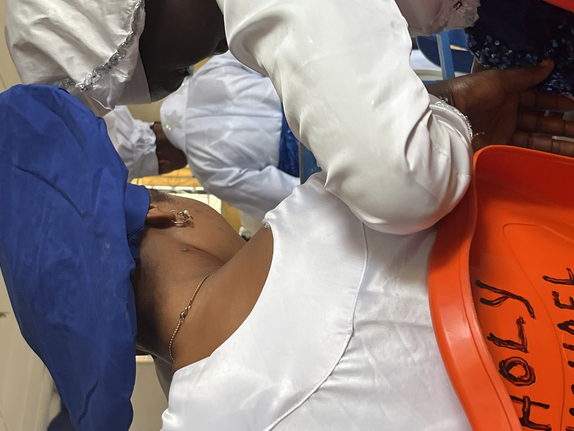 The image shows the back and side of three individuals wearing white garments and headwear, likely in a religious setting. The person in the foreground on the left has a blue head covering and is wearing a white garment with a gold chain and earrings. To their right, another person in a white garment is partially visible, with their arm resting on the orange seat in front of them. A third person, seen from behind, is wearing a white garment and a white head covering with silver trim. In the background, another individual in a white garment with a blue sash is visible. The orange seat has handwritten text in black marker reading "HOLY MICHAEL". The environment appears to be indoors, possibly a place of worship or a communal gathering. The lighting suggests it is daytime.