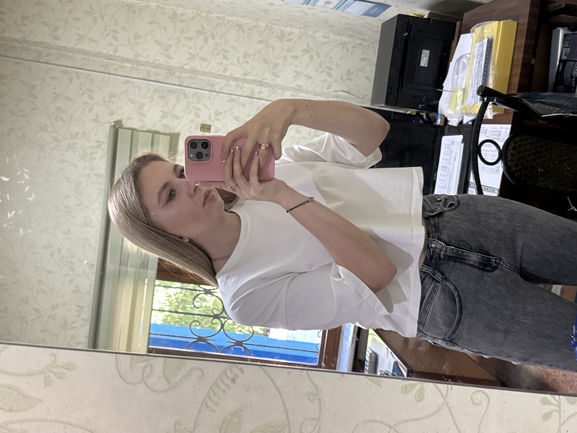 A young woman with blonde hair takes a mirror selfie in an indoor setting. She is wearing a white cropped t-shirt and dark grey jeans. She holds a pink cell phone in her right hand, which is angled to capture her reflection. A ring is visible on her left ring finger, and a bracelet is on her left wrist. The background includes a mirror reflecting a window with bars and greenery outside, a desk with papers and a printer, and patterned wallpaper.