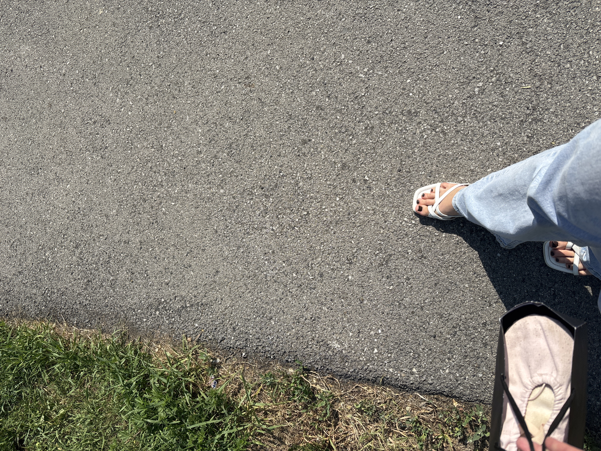 Inskoy, Russia. A person is walking on an asphalt path next to a patch of grass. The person is wearing light blue jeans and white sandals. They are carrying a dark shopping bag in their left hand, which contains a light-colored item. The sun is casting a shadow from the person onto the asphalt. The scene is outdoors, with the asphalt path appearing to be a sidewalk or walking trail.