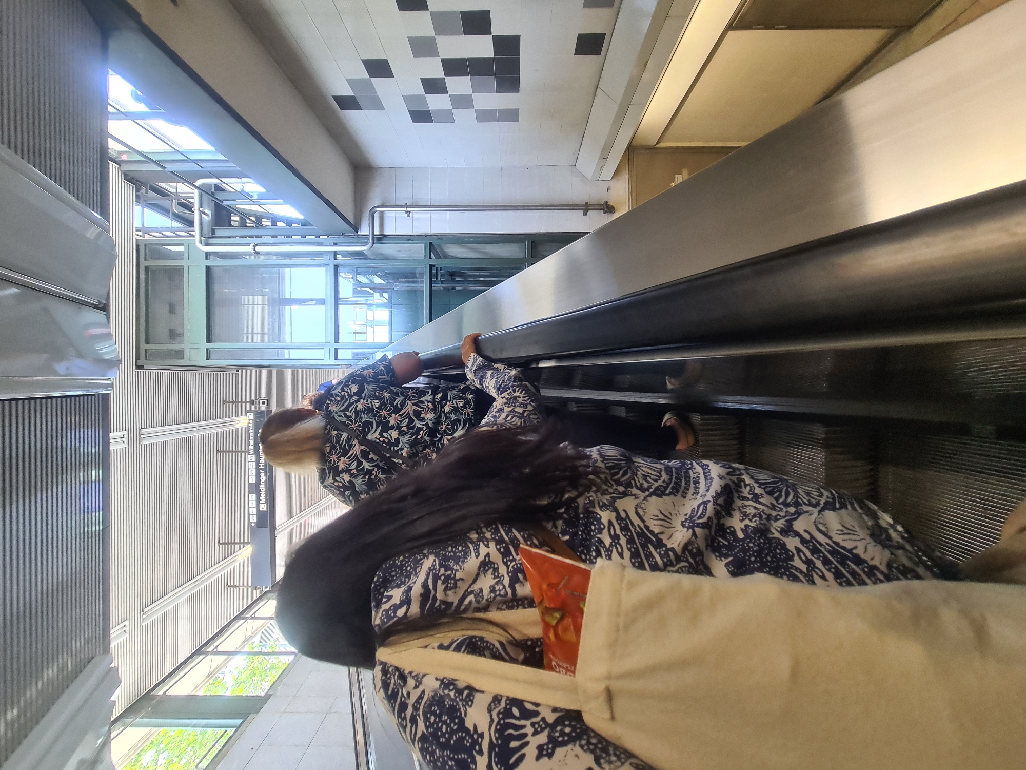 Two individuals are ascending an escalator within a public transit station in Vienna, Austria. The person in the foreground, facing away from the camera, has long dark hair and is carrying a light-colored tote bag on their left shoulder. They are wearing a dark blue and white patterned shirt. Behind them, another person with blonde hair, also facing away, is wearing a similar dark blue and white patterned shirt. The escalator is moving upwards, indicated by the inclined steps. Above the escalator, a metal ceiling with fluorescent lighting is visible. To the left, there are windows looking out onto an area with trees and greenery, suggesting an outdoor environment. A sign with German text, "Meidlinger Hauptstraße" and "Wilhelmstraße," is visible overhead, indicating the location as a transit hub in Vienna. To the right, a glass elevator shaft with metal railings and stairs is present. The wall to the right of the escalator is tiled with a mosaic pattern of light and dark gray squares. Below the escalator, a recessed area with beige-colored walls and a doorway is visible. The overall environment appears to be a modern and well-maintained urban transit facility.