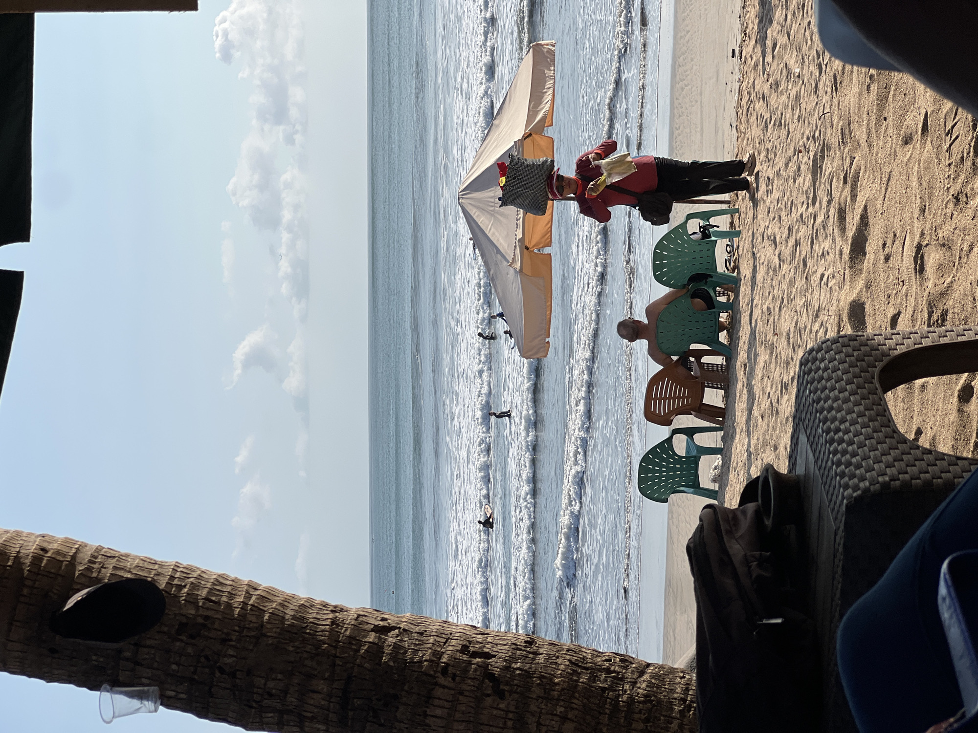 The image depicts a tropical beach scene in Kuta, Indonesia, with a prominent palm tree trunk in the foreground on the left. Several people are visible, including surfers in the ocean and individuals relaxing on the beach. A vendor, wearing a red shirt and dark pants, is standing under a beach umbrella, holding what appears to be food items. Two people are seated on chairs facing the ocean, with a third person behind them. Beach chairs and a table are present on the sand. The ocean features moderate waves, and there are scattered clouds in the sky.