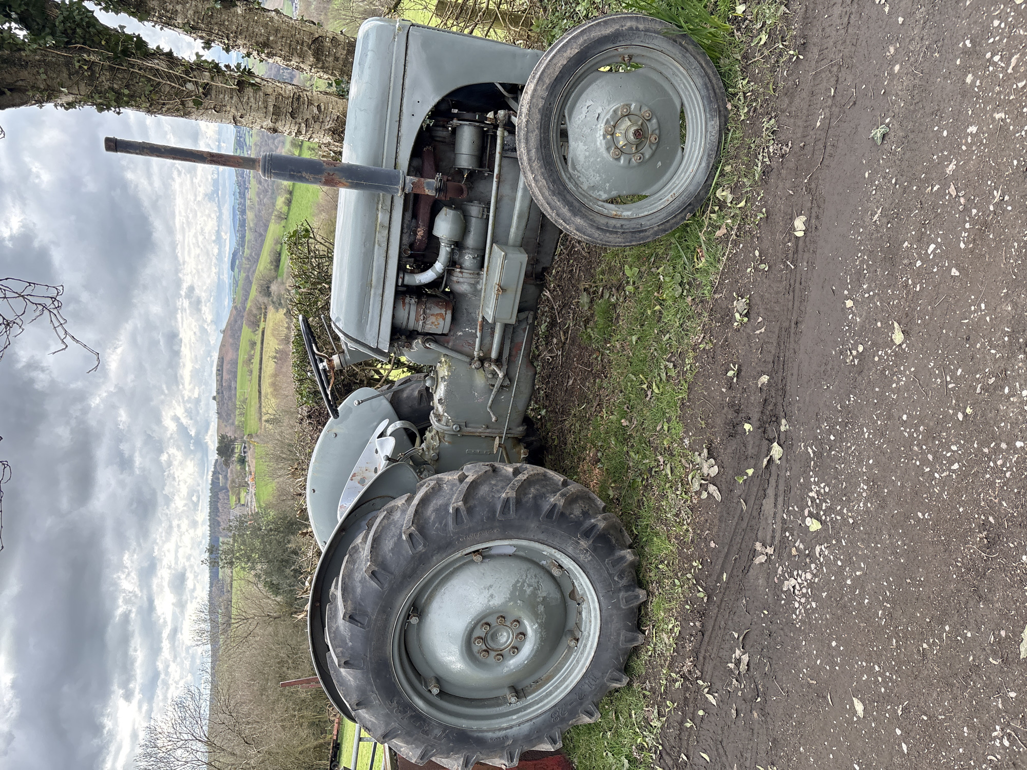 A gray, vintage tractor is parked on a grassy verge adjacent to a dirt road in Stiperstones, United Kingdom. The tractor is viewed from the side, with its large rear tire prominent and a smaller front tire also visible. The engine and exhaust pipe are detailed, exhibiting signs of age and rust. The background shows a landscape of green fields and distant hills under a cloudy sky. Some trees and hedges border the field, adding depth to the scene.