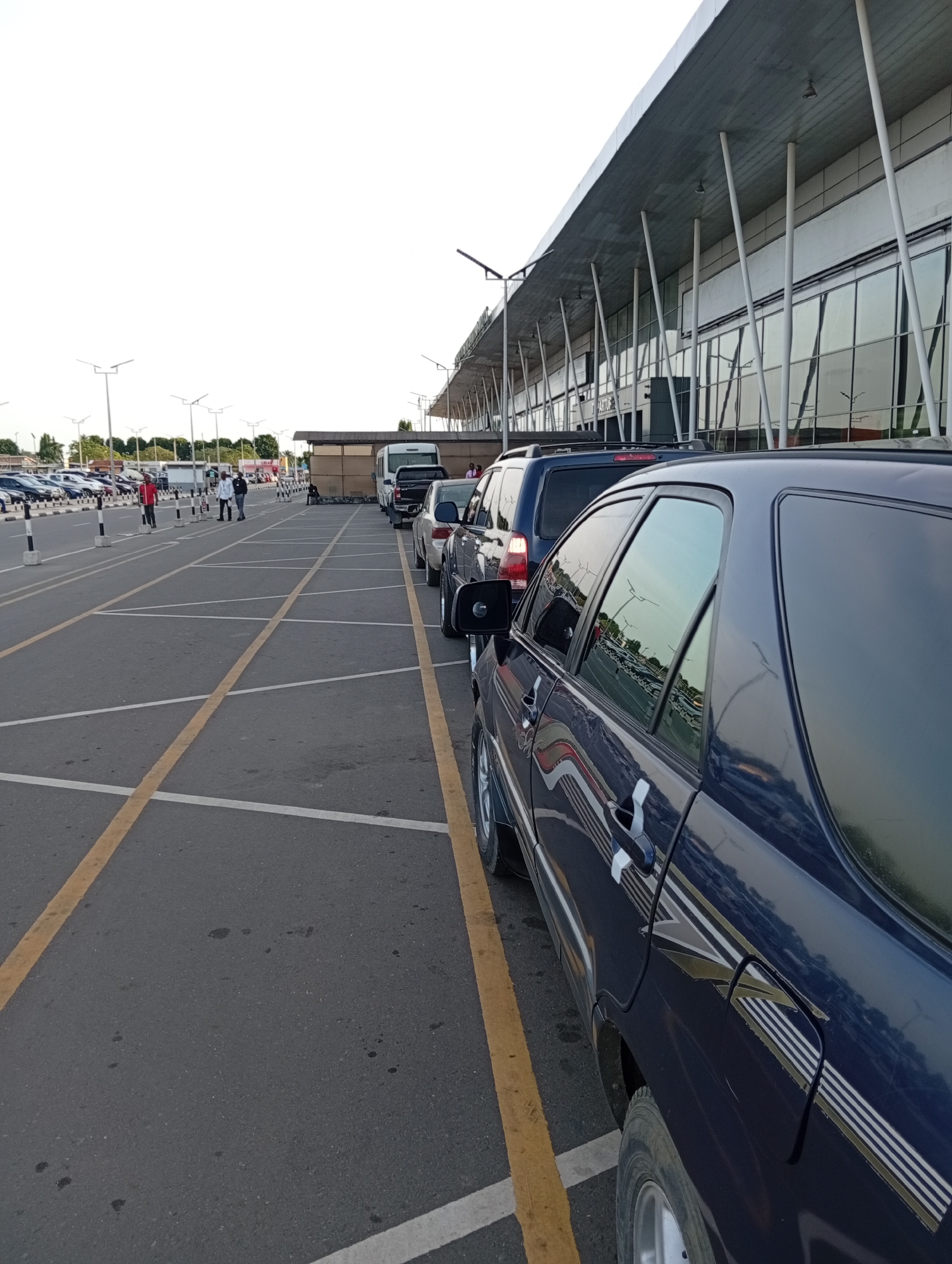 The image depicts a parking area in Omagwa, Nigeria. Parked cars line the right side, with a dark blue car in the foreground. The parking lot has yellow lines marking parking spaces, with white lines dividing rows. In the background, people stand near a terminal building with a modern design, featuring large windows and supporting columns. Light poles are visible throughout the parking area. Other vehicles, including a white van and several cars, are parked further back. The sky is overcast.