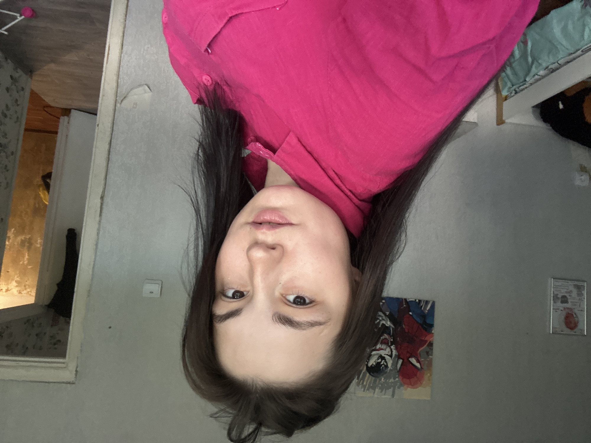 A young woman with dark hair and brown eyes is taking a selfie in a room. She is wearing a bright pink collared shirt. Behind her, on the wall, are two framed pictures, one of Spider-Man and Venom, and another smaller framed item to the left. To the right, a doorway leads to another room with patterned wallpaper and a doorway into what appears to be another room. The walls of the room she is in are light gray, and there is a light switch on the wall. To her left, a white bed frame with bedding is visible. The image is likely taken indoors in an urban environment, possibly an apartment in the city of Chaikovsky, Russia.