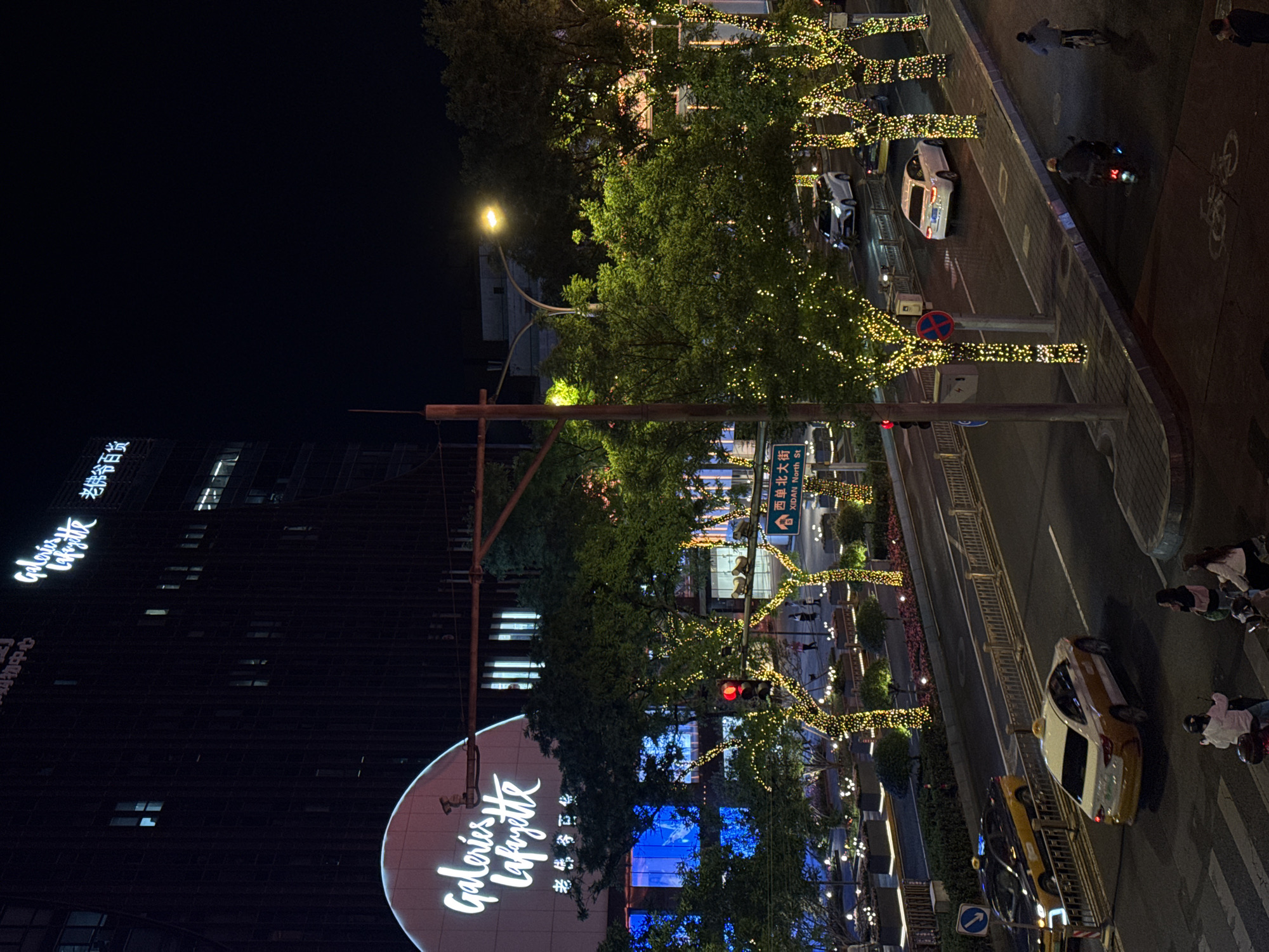 The image, taken at night in Xicheng District, China, showcases a cityscape view. A tall building, identified by "Galeries Lafayette" signage in both English and Chinese, dominates the left side. A street scene is visible on the right, featuring trees adorned with strings of lights. Cars, including a taxi, travel along the street, and pedestrians are also present. Street signs indicate the location as "XIDAN North St." The overall impression is that of a bustling urban environment at night.