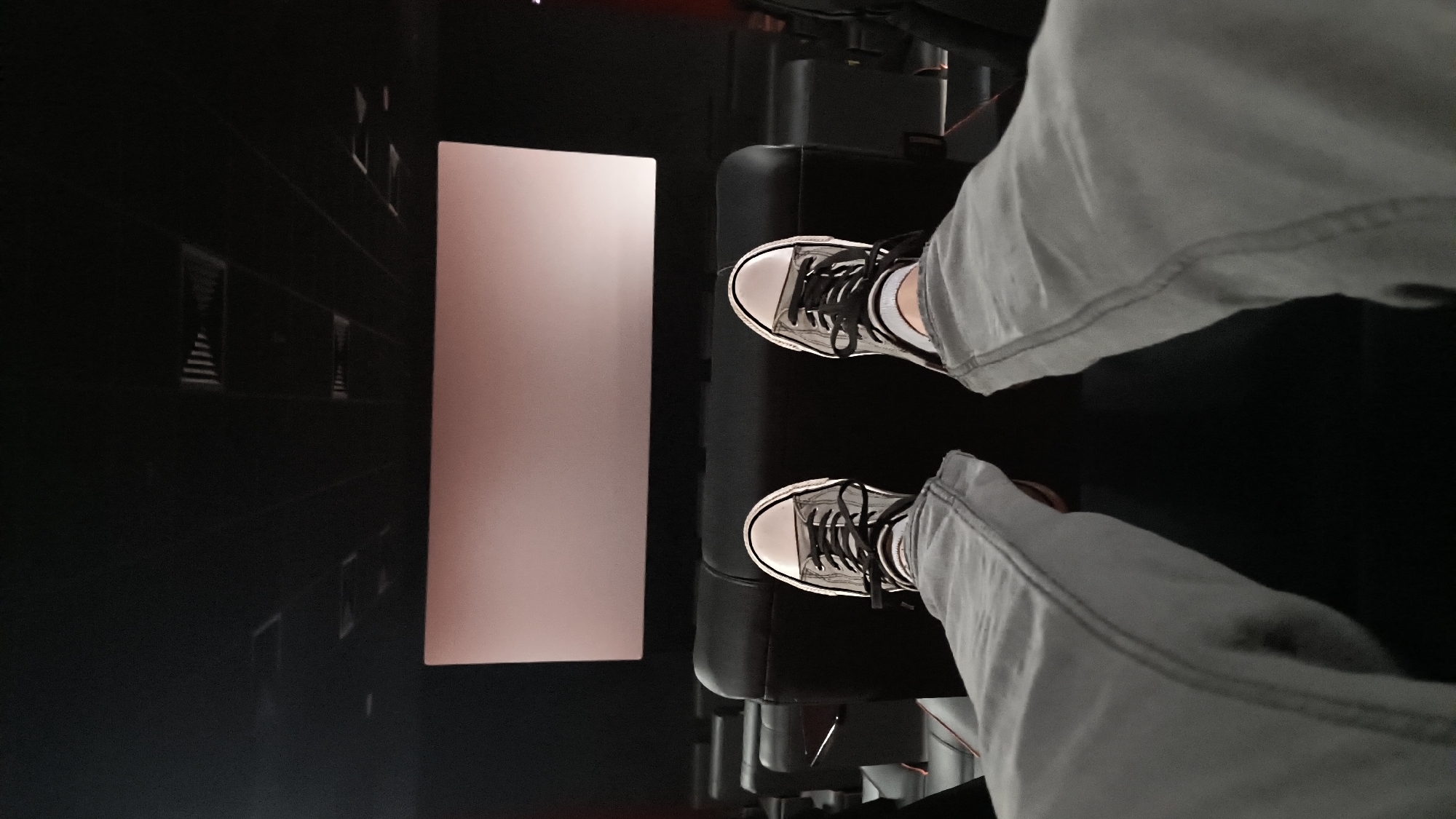 The image depicts a first-person perspective from a seated position, likely in a motion simulator or similar experience. The viewer is wearing light-colored jeans and black and white sneakers. In front of the viewer is a rectangular screen displaying a pinkish-white color. To the left of the screen, there is an image of a dark, urban environment with repeating architectural elements that could be buildings. The viewer is seated in a black chair. The scene appears to be indoors in a darkened room. There is no identifiable information to ascertain if the location is in Sofia, Bulgaria.