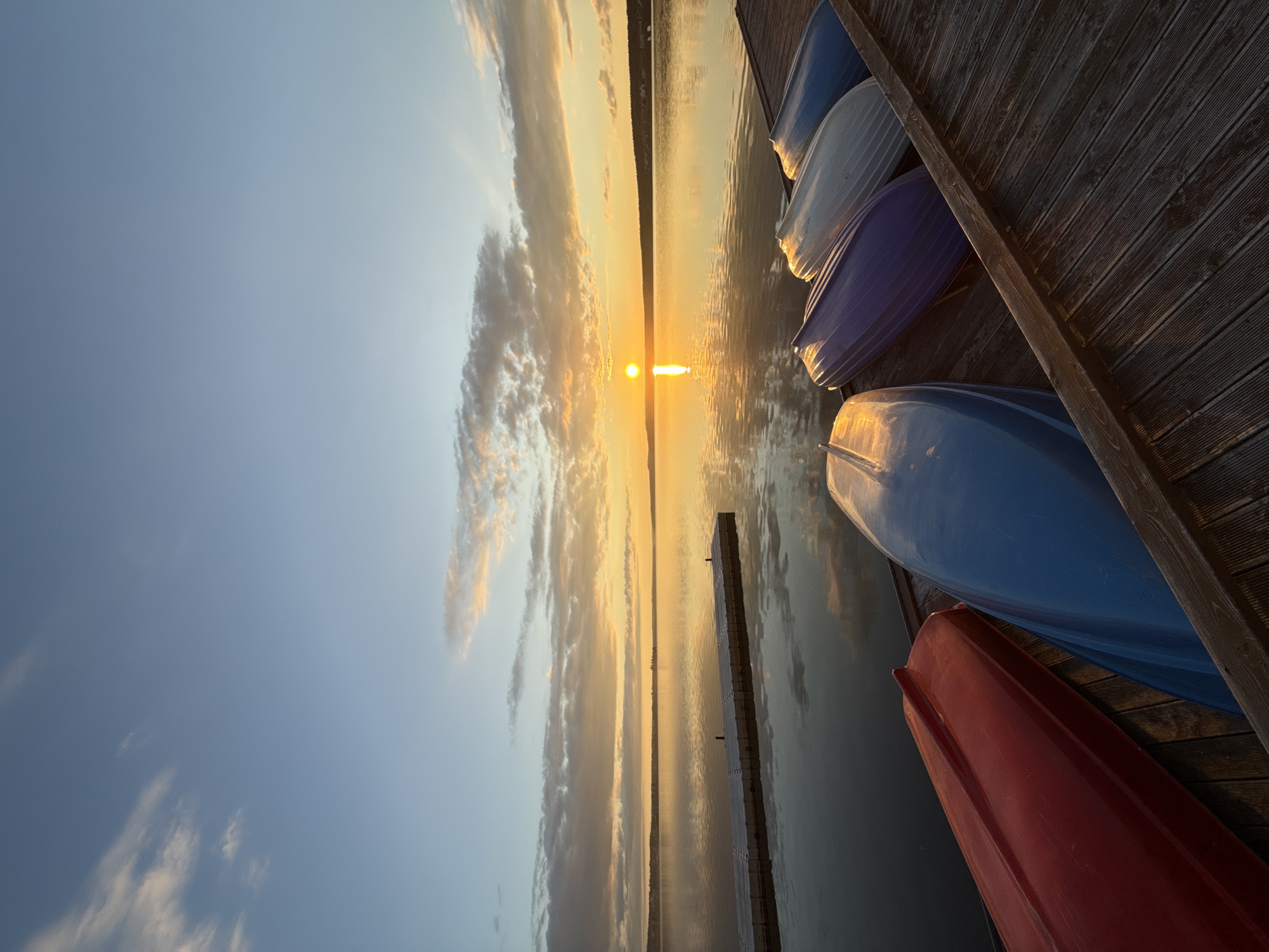 This image captures a serene sunrise or sunset over a wide body of water, likely a lake, in Votkinsk, Russia. The sky, occupying the upper portion of the frame, transitions from a clear blue at the top to warm golden and orange hues near the horizon. Wispy clouds are scattered across the sky, glowing with the reflected light.
A bright, circular sun is positioned low on the horizon, creating a striking vertical path of brilliant reflection across the calm water. The water's surface, smooth and still, mirrors the vibrant colors of the sky and the sun's glow. A distant, dark landmass, possibly a tree-lined shore, forms a subtle silhouette along the far horizon.
In the foreground, a sturdy wooden dock extends along the bottom and right edges of the frame. Several small boats, possibly kayaks or rowboats, are stored upside down on this dock. Their undersides are visible in various colors, including blue, white, purple, and red, all bathed in the warm, golden light of the low sun. Another narrower wooden pier extends further into the water from the left foreground, creating leading lines towards the center of the peaceful scene.
The image conveys a tranquil atmosphere, emphasizing the natural beauty of the golden hour. There is no human activity depicted, only the quiet presence of the boats waiting on the dock. No visible text is present.