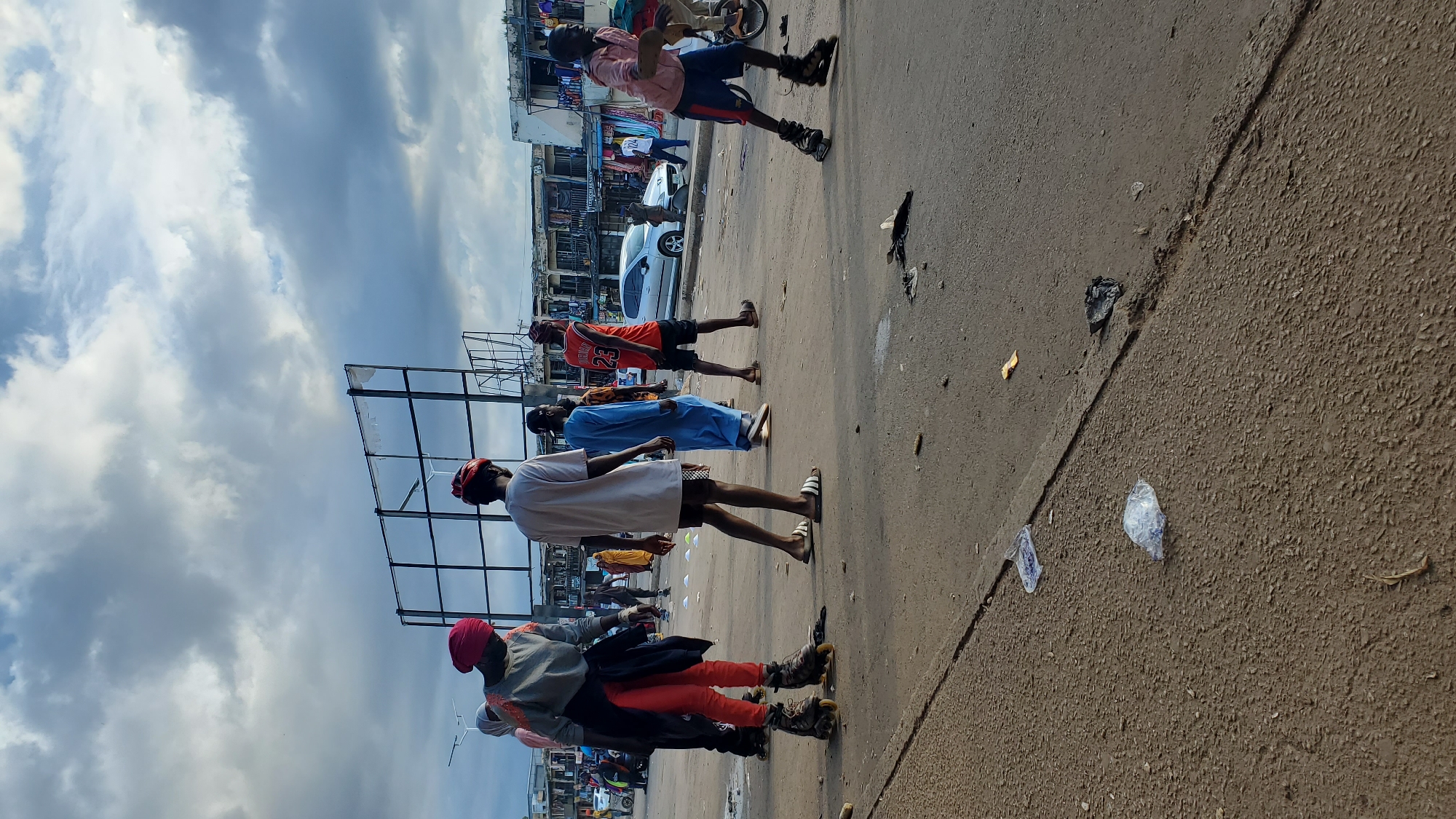 The image depicts an outdoor scene in what appears to be a city. In the foreground, the asphalt road shows signs of debris, including plastic bags and wrappers. Several individuals are present, some engaged in rollerblading. One person on the left wears bright orange pants and a red headscarf, rollerblading. Another individual in the center wears a white t-shirt and shorts, walking. To the right, a person in a red basketball jersey with the number 23 is also walking, and further to the right, another person is rollerblading in a pink shirt and blue shorts. In the background, buildings line the street, some appearing to be shops with merchandise displayed. A billboard structure is visible on the left, and a silver car is parked in the middle ground. The sky is overcast with dramatic cloud formations. The overall impression is a lively, urban environment with people participating in recreational activities on the street.