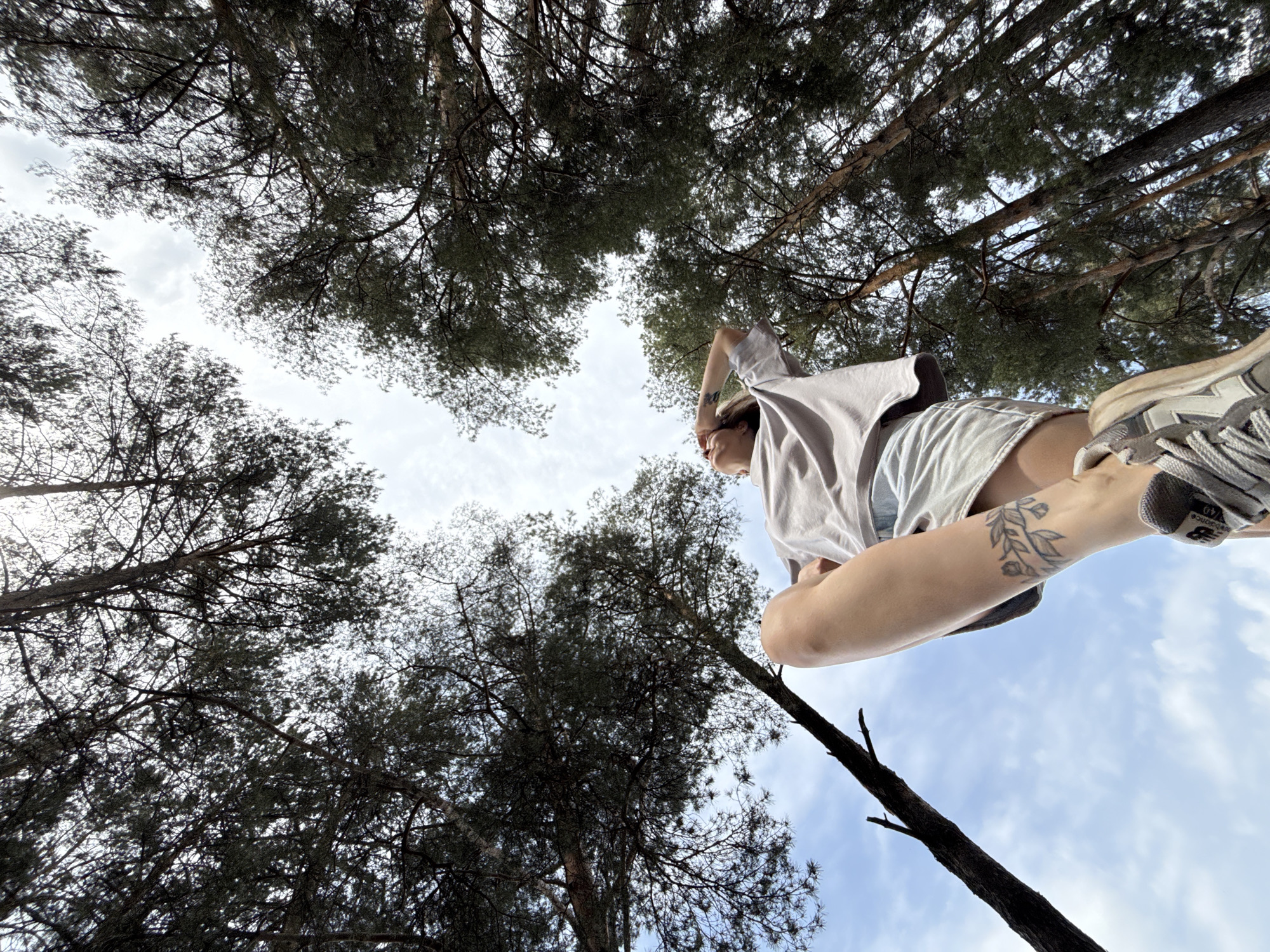 The image is a low-angle shot looking up through a canopy of coniferous trees. A person is in the foreground, seen from the feet up. They are wearing shorts, a t-shirt, and sneakers. A floral tattoo is visible on their leg. The location is likely a forest or park in Chaikovsky, Russia, given the prompt. The sky is visible through gaps in the tree branches.