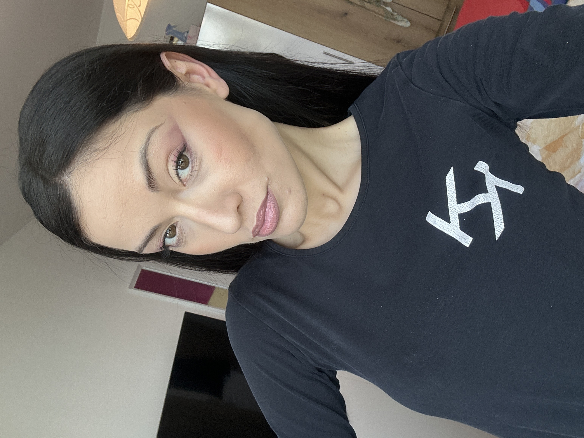A person with long, dark hair and fair skin is seen from a close-up, tilted perspective. They are wearing a black long-sleeved shirt with a white logo on the front. The person's makeup includes eyeshadow and lipstick.
The background is a mix of domestic interior elements, including a light-colored wall, furniture, and a television screen. The scene seems to be indoors. There is no definitive information linking the location to Burgas, Bulgaria.