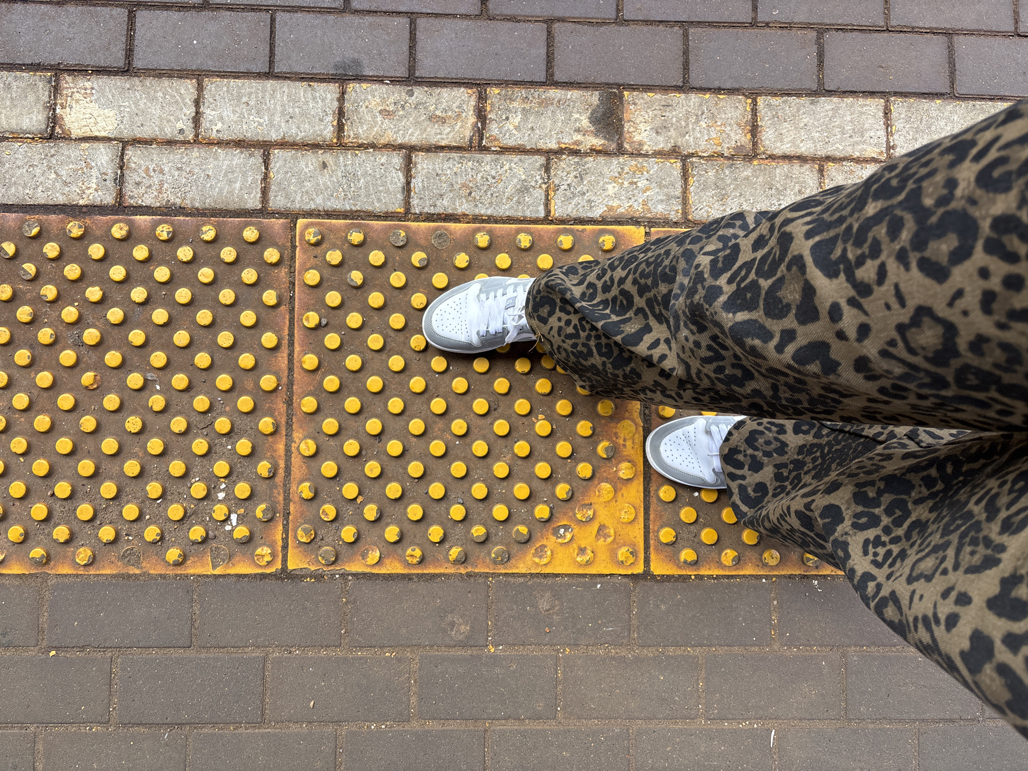 The image shows a top-down view of a person standing on a tactile paving surface, likely at a transportation hub. The paving consists of yellow circular bumps on a darker background, signaling a change in surface or a potential hazard. The person is wearing white sneakers and patterned pants. The tactile paving is bordered by brickwork, suggesting a sidewalk or platform. The location is Moscow, Russia.