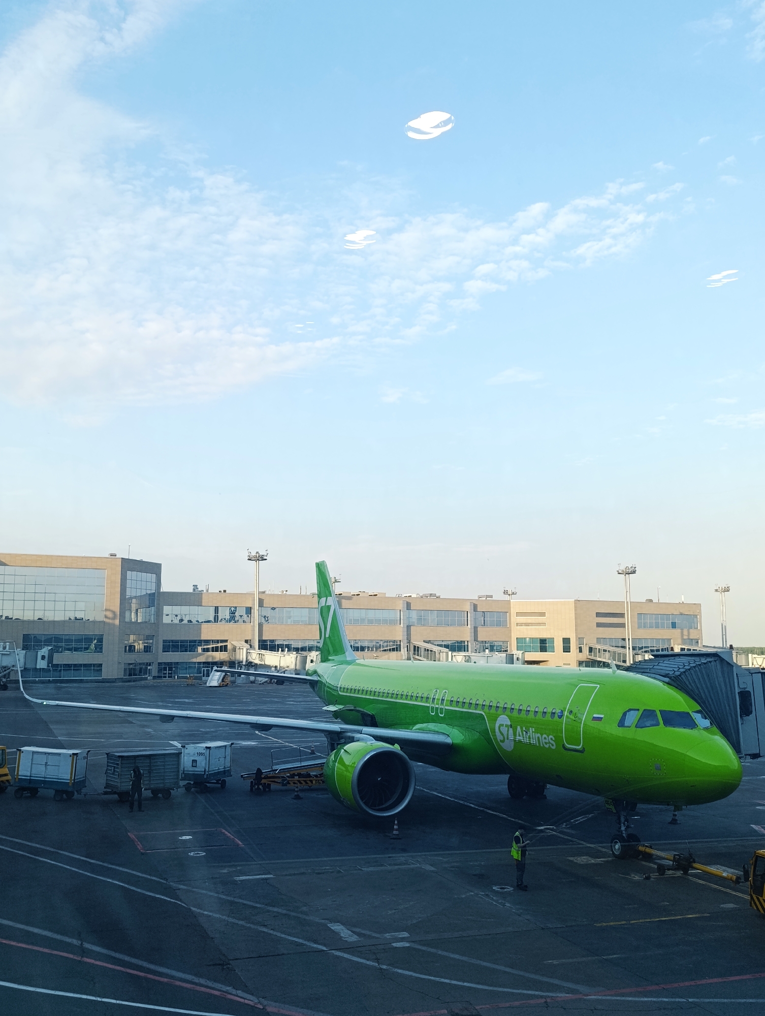 This media file captures an airplane at an airport gate on a clear day. The primary subject is a bright green S7 Airlines passenger jet, parked next to a modern airport terminal building. The plane is positioned at an angle, with its nose pointed towards the viewer's right. The S7 Airlines logo is prominently displayed on the fuselage, along with the text "S7 Airlines".
In the foreground, on the tarmac, several airport service vehicles and cargo containers are visible. One person, wearing a high-visibility vest, stands near the front of the airplane, seemingly overseeing operations. Another individual in a similar vest is seen further back, near some cargo containers. The airport terminal features large glass windows, reflecting the sky. The overall scene suggests a typical airport operation, possibly preparing for departure or having recently arrived.
The sky is a clear blue with scattered, wispy white clouds, indicating daytime, likely mid-morning or afternoon. The lighting suggests bright sunshine. The presence of the airport terminal, jet bridge, and ground support equipment firmly places the setting at an airport. Given the "S7 Airlines" branding, the location is likely an airport served by this Russian airline, with Domodedovo, Russia being the specified context. There are no strong emotional cues, but the scene conveys a sense of organized activity. Small white, ring-like shapes are visible in the sky, which might be lens flare or reflections.