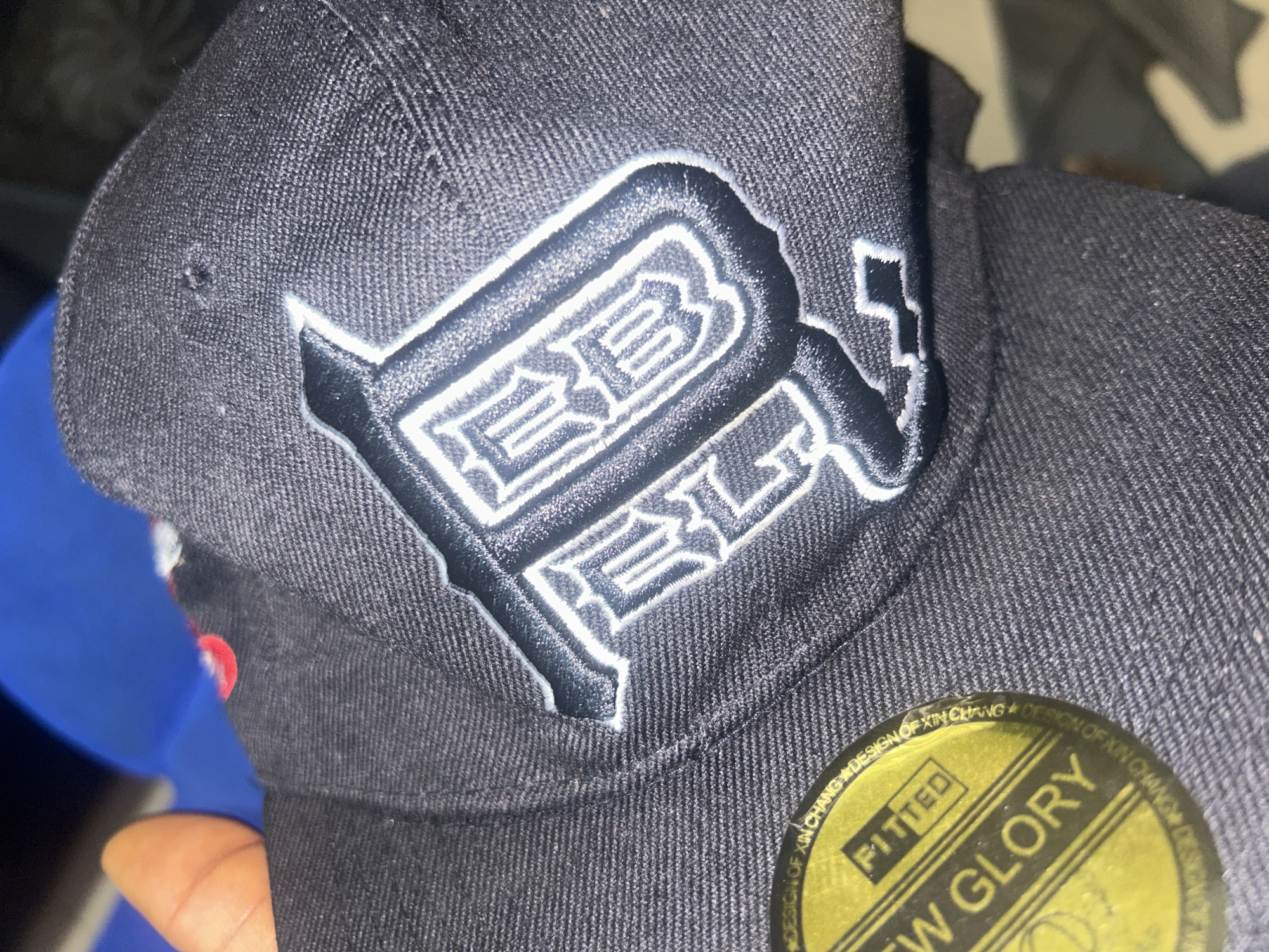 The image shows a close-up of a black fitted baseball cap with a white and black embroidered logo. The logo appears to be stylized lettering. A yellow sticker with the words "FITTED" and "NEW GLORY" is visible on the brim of the cap. The background is blurred and shows a hint of a blue object. The lighting suggests an indoor setting. There is no indication of the city of Ogbomosho, Nigeria in the image or its surrounding context.
