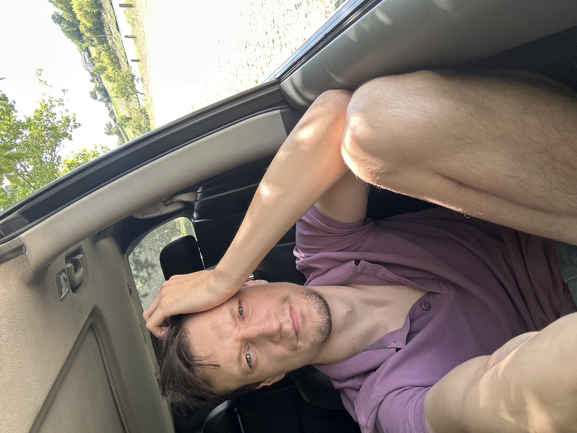 In Makiv, Ukraine, a man is pictured inside a car, seated with his left arm resting on his head. He is wearing a purple polo shirt and has short brown hair and a stubble beard. Sunlight streams into the car, illuminating the scene. Outside the car, a rural landscape is visible, featuring a river, trees, and houses on a hill. The image appears to be a selfie taken by the man.
