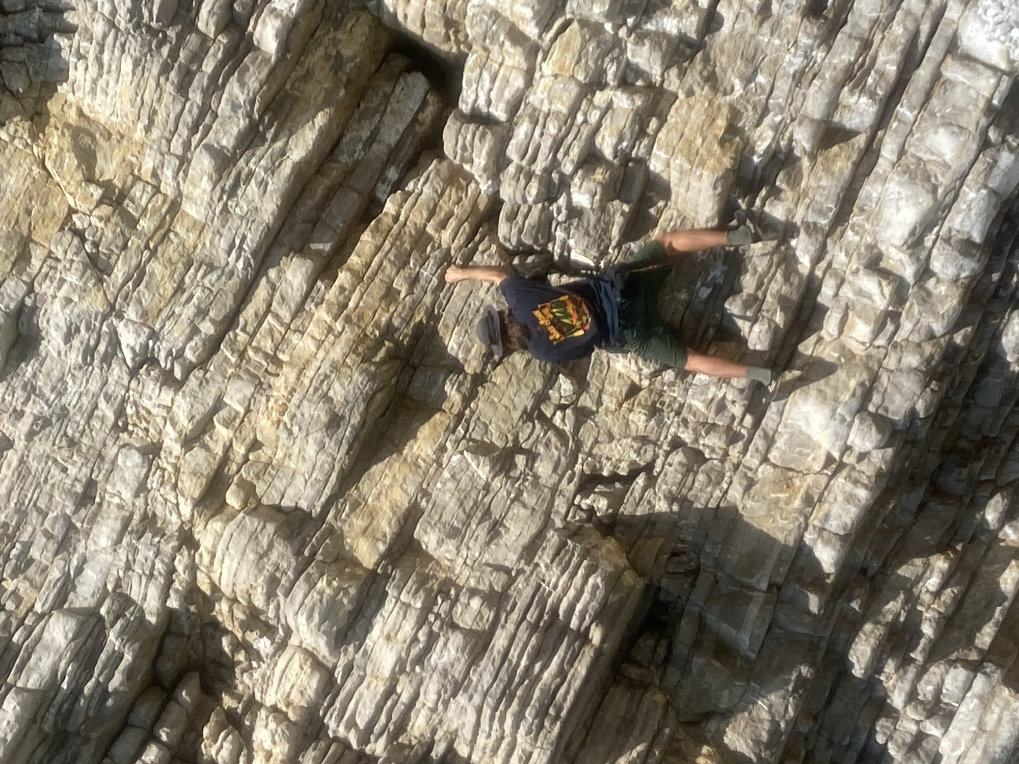 A person is spread-eagled on a rock face, seemingly engaging in a climbing activity. The person is wearing a dark-colored t-shirt with a graphic design on the back, olive green shorts, and a baseball cap. They are positioned diagonally across the frame. The rock face is textured, with layered geological formations, predominantly in shades of gray and beige. The overall perspective is from above, as if the image was taken from a drone or an elevated vantage point. The scene is bathed in sunlight, creating stark shadows that accentuate the rock's topography. The location is Budva, Montenegro, specifically on a rocky coastal area or cliff face.