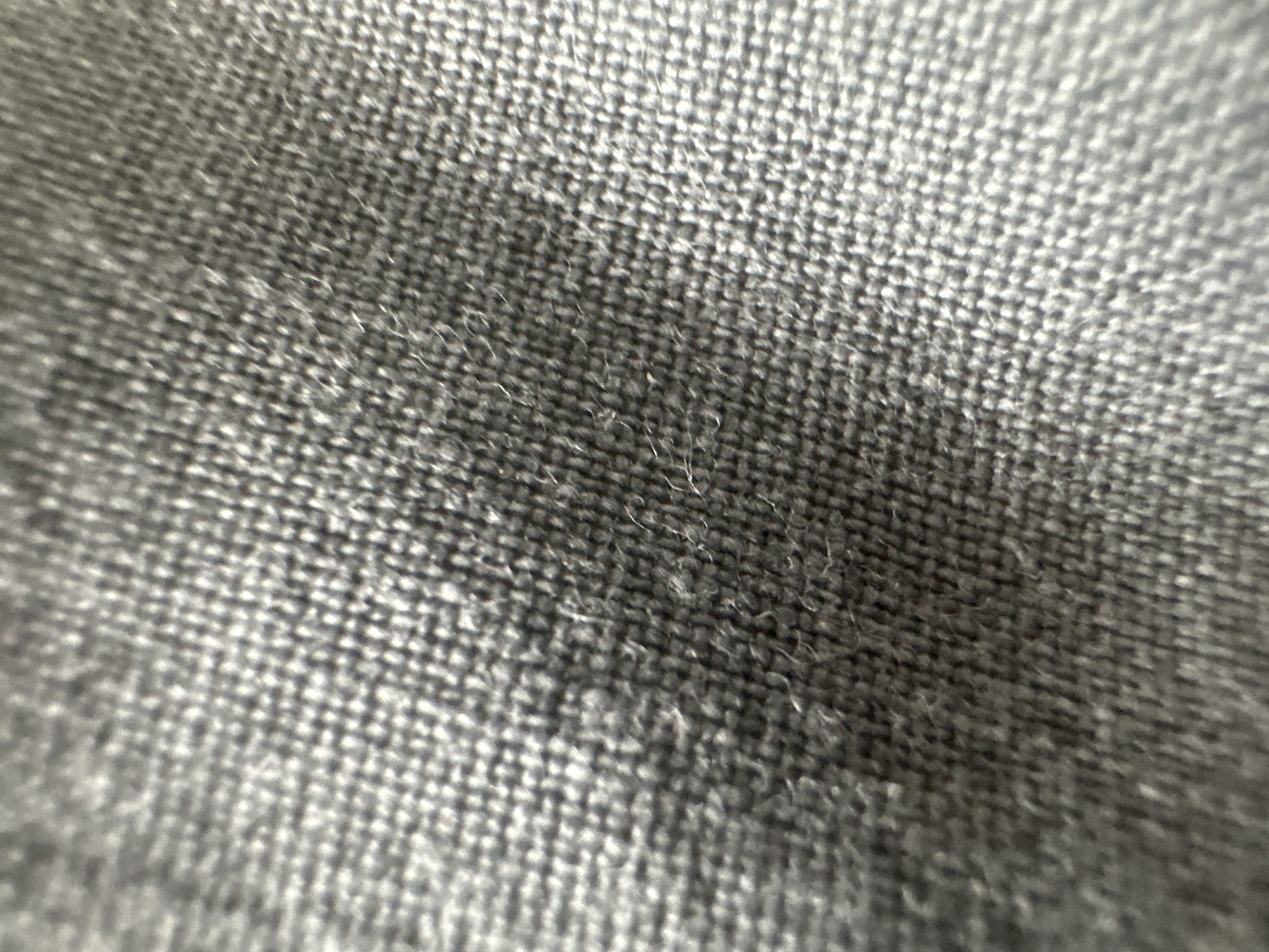 This is a macro photograph of a grey, woven fabric. The texture of the weave is clearly visible, with individual threads forming a grid-like pattern. Some stray fibers are also apparent, giving the fabric a slightly fuzzy appearance. The lighting creates highlights and shadows that emphasize the texture. There are no people, objects, or environmental features discernible in this image; it is solely focused on the material's surface. The image is not from Belgrade, Serbia.