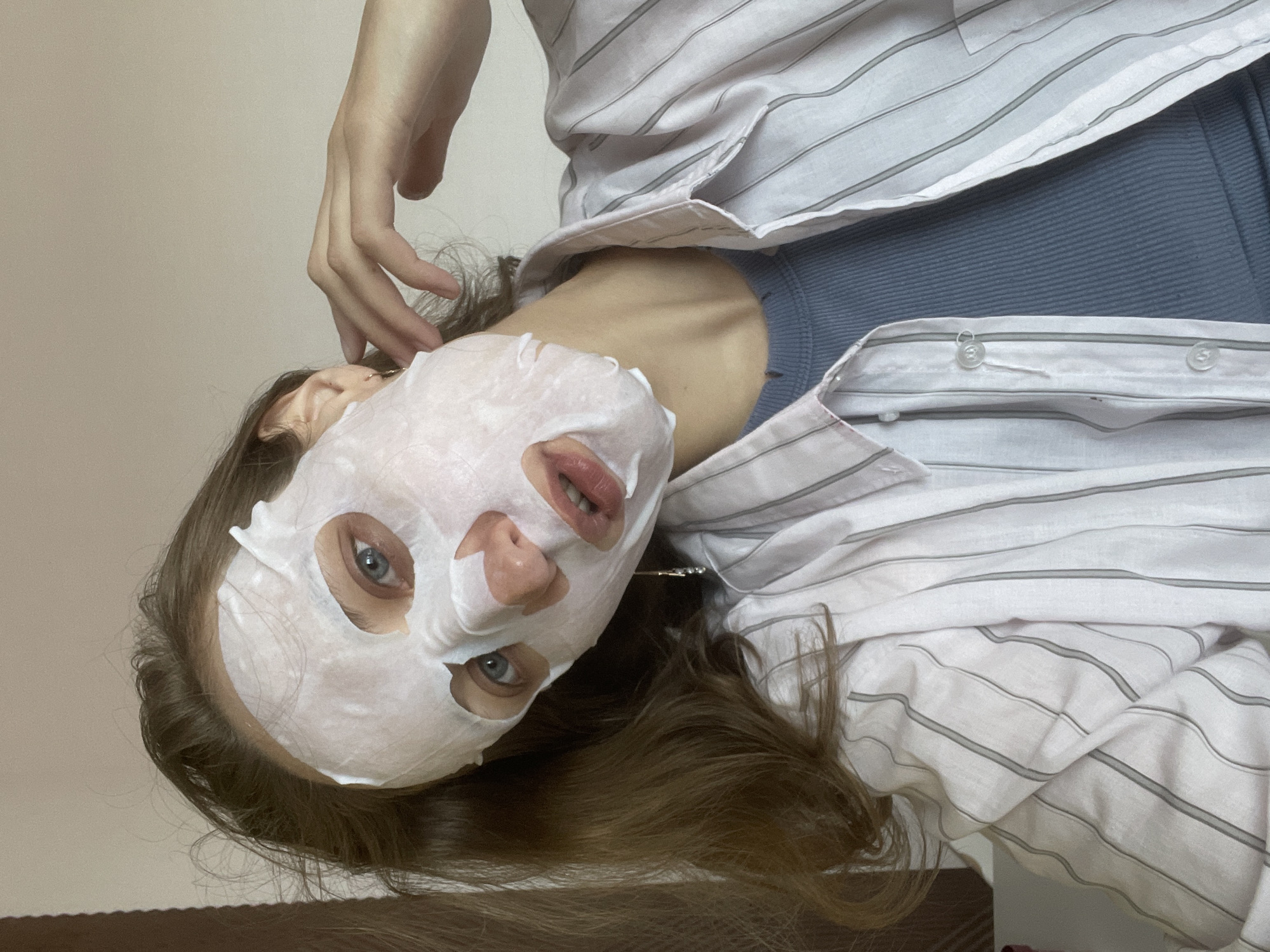 A woman with a white sheet mask is shown. She is wearing a white and gray striped button-down shirt and blue ribbed tank top. Her left hand touches her hair. Her long brown hair flows down. The location may be Sepych, Russia, but there is no evidence within the image.