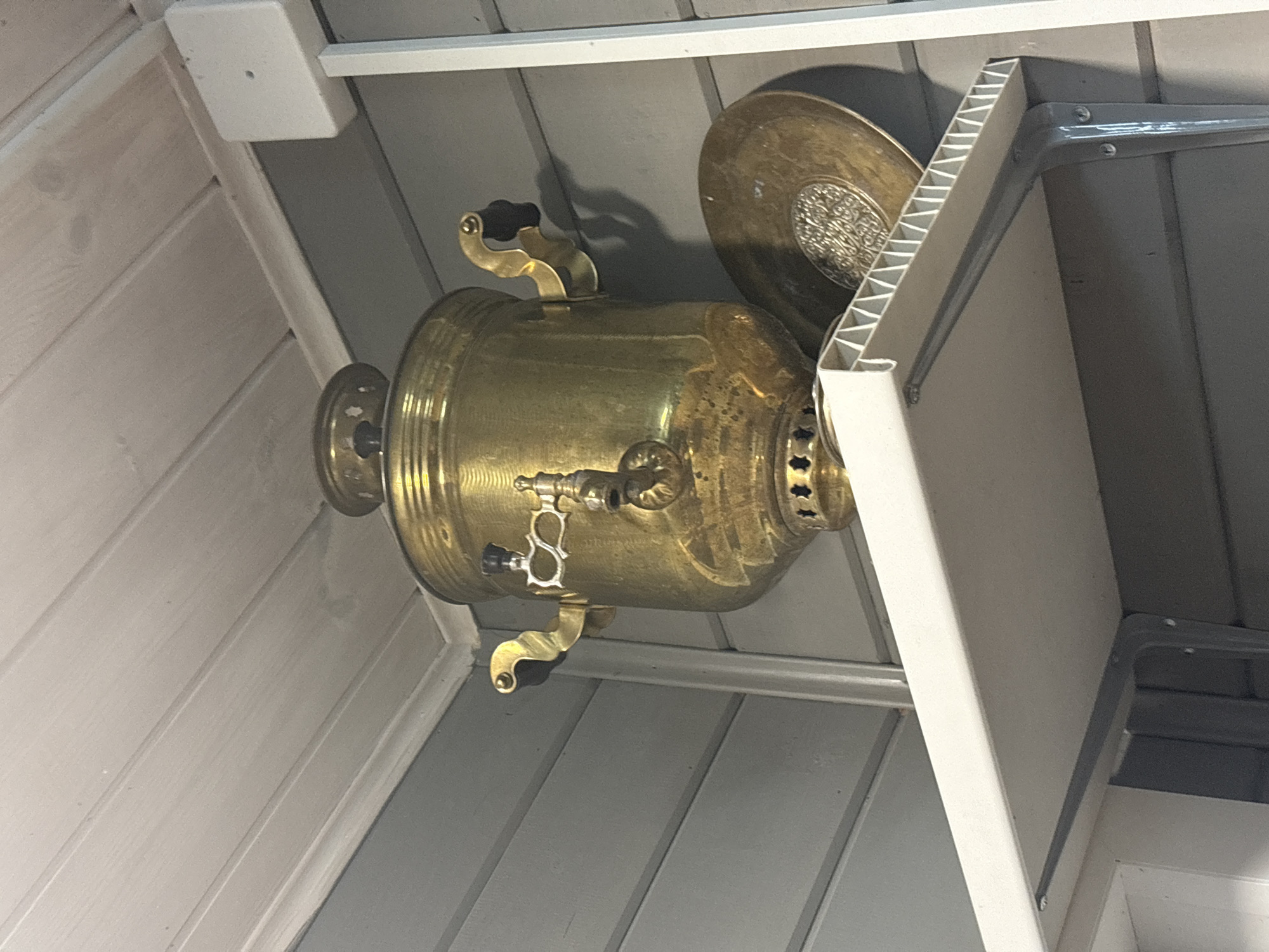 A brass samovar sits on a white shelf, to the right of it is a brass plate. The background is a grey, horizontal wooden panel wall with a white wooden ceiling above. The scene is indoors, likely in a storage area or a rustic room. The lighting is dim, suggesting it might be evening or the area is not well-lit. There are no people visible in the image. The overall impression is of a still life, with the samovar and plate being the focal points.