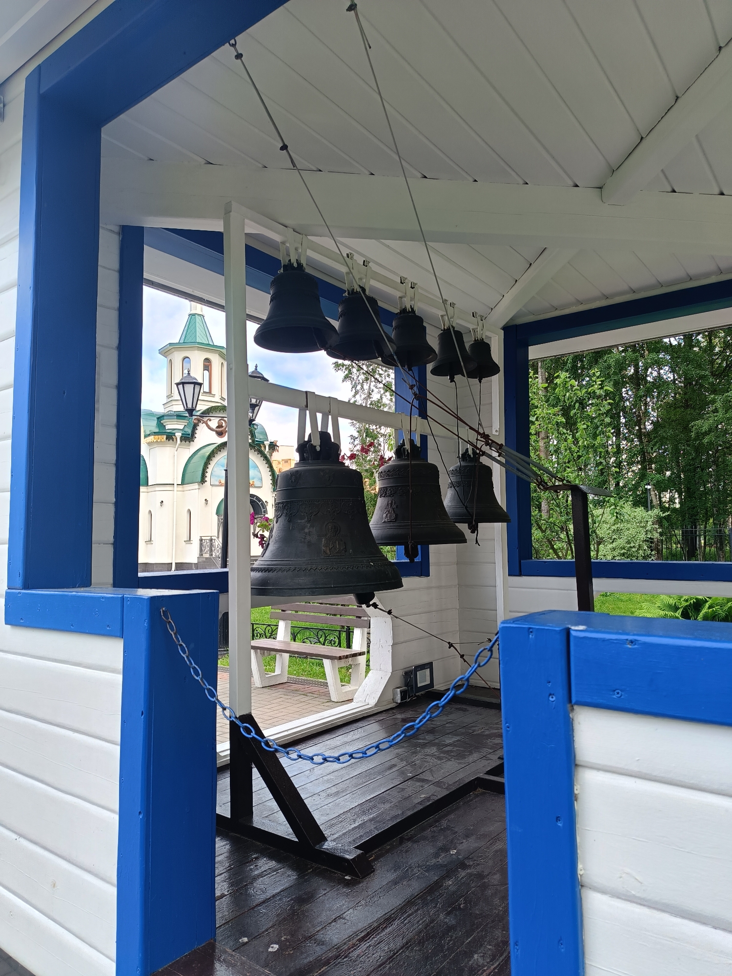 A set of bells hangs from a white wooden structure with blue trim. The structure is open on one side, revealing a church with green domes in the background, a wooden bench, and lush green trees. The ground beneath the bells is made of dark wooden planks, and a blue chain is strung across the entrance. The scene is peaceful and appears to be outdoors.