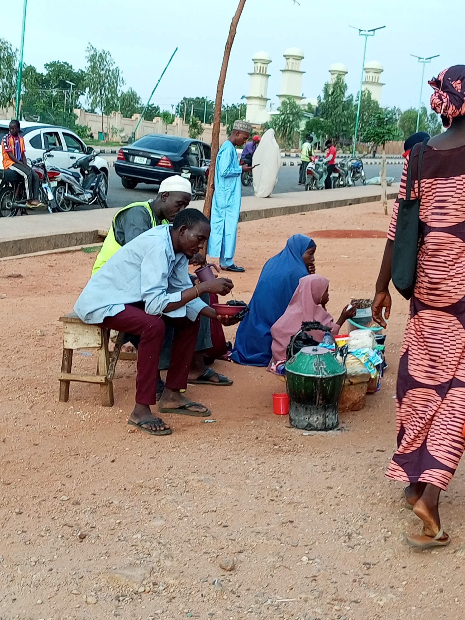 The image depicts a street scene in Gombe, Nigeria, with a group of people gathered. In the foreground, two men are seated on a wooden stool, one of whom is drinking from a cup. To their right, two women are kneeling on the ground, one in a blue hijab and the other in a pink hijab. They are surrounded by various containers and items, suggesting they are vendors. Behind them, other individuals and motorcycles are visible, and in the background, modern white structures with domes, resembling mosque minarets, are prominent. The ground is covered in dirt and gravel.