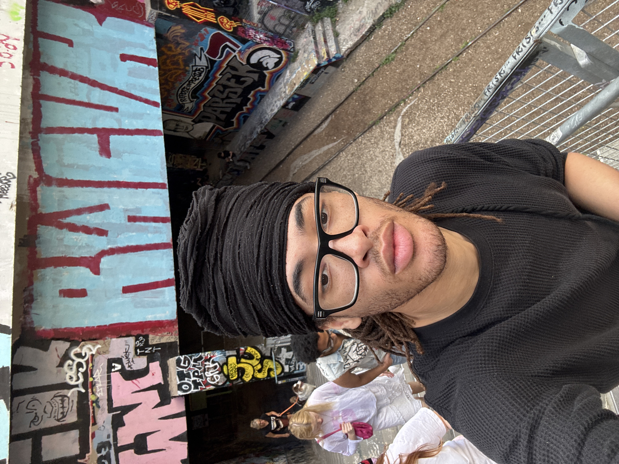 A selfie of a person with dreadlocks and glasses, wearing a black shirt and head covering, is oriented vertically. The person appears to be on a walkway overlooking a lower level with graffiti-covered walls. A group of people, some wearing white clothing, are walking below. The location is likely in Paris, France, due to the graffiti style and urban setting.