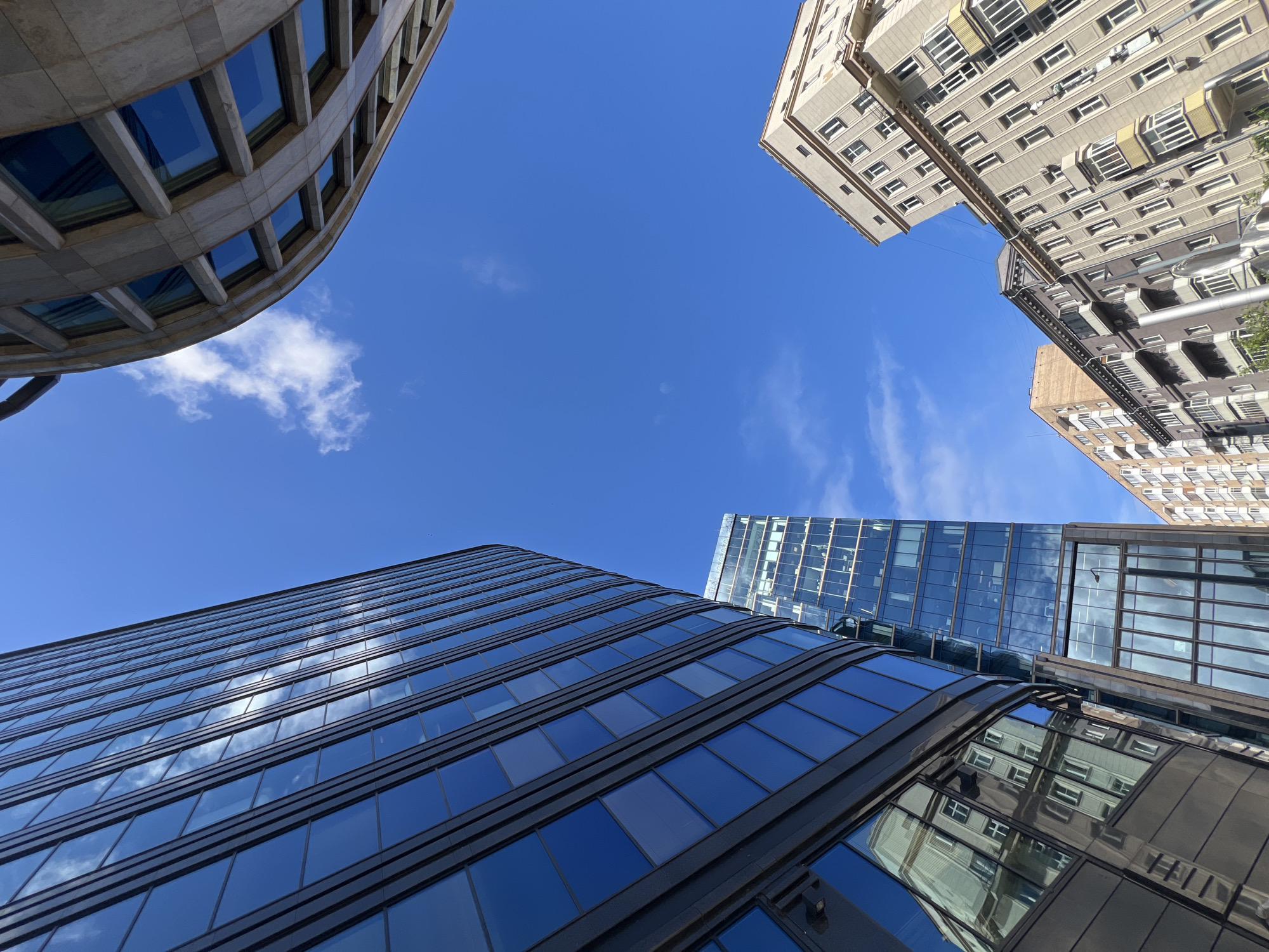 The image shows a low-angle view of several buildings in Moscow, Russia, against a clear blue sky with scattered clouds. The architectural styles vary, with one building featuring a curved facade and rectangular windows, and another with a grid-like pattern of glass windows reflecting the sky. Two older buildings, are visible on the upper right corner.