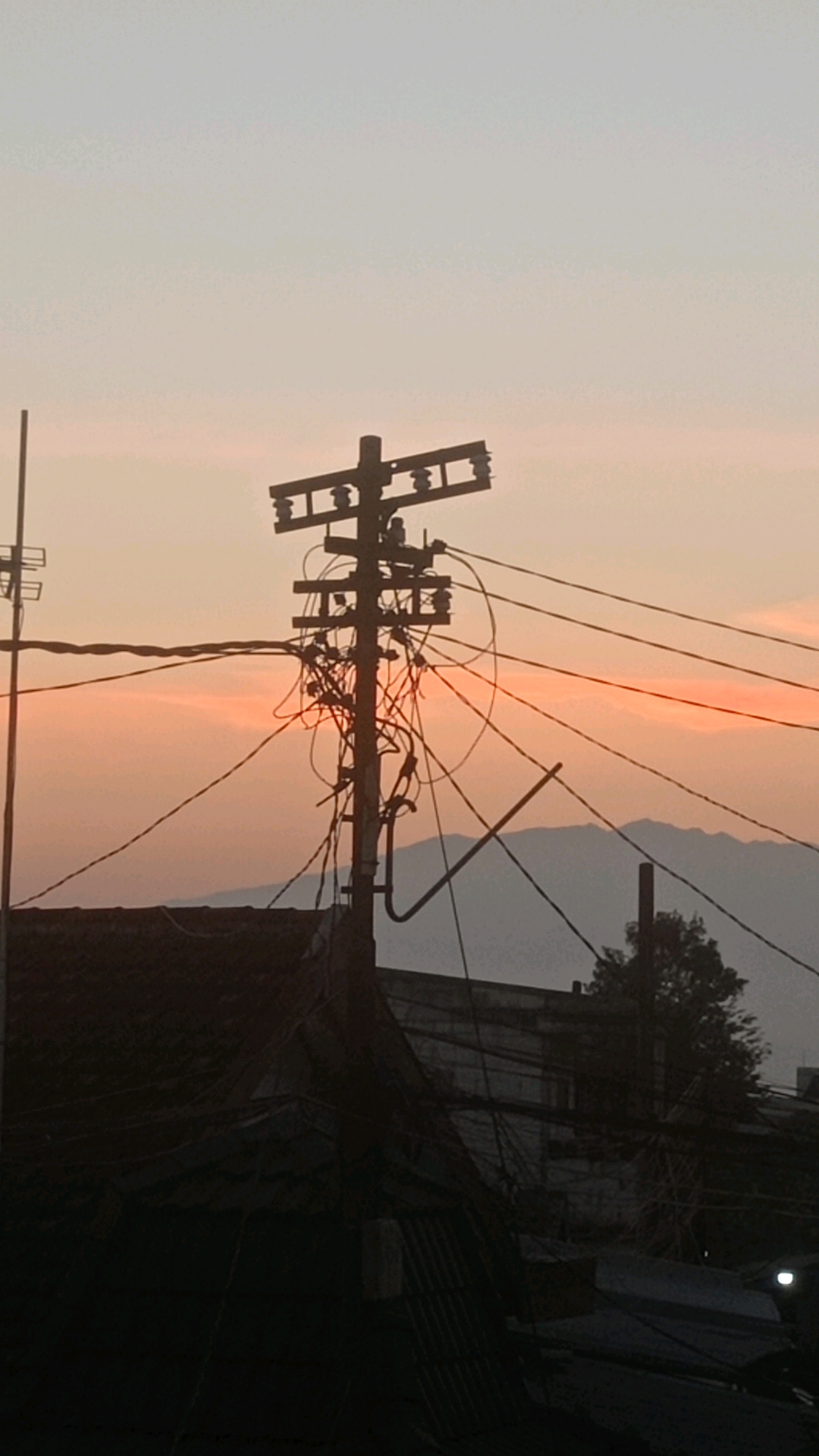 A silhouette of a utility pole with tangled wires is visible against a hazy, peach-colored sky, suggesting dawn or dusk. The pole stands in front of a backdrop of dark, low-lying buildings and distant mountains. The sky is lighter at the top and graduates to a warmer hue near the horizon, with faint streaks of pink and orange indicating the presence of the sun. The utility pole is heavily laden with a complex network of cables, appearing disorganized and chaotic. Below the pole, the roofs of several buildings are silhouetted against the sky, their textures appearing corrugated or metallic in places. The overall scene is characterized by muted colors and a somewhat dystopian aesthetic, with the dense web of wires dominating the composition and obscuring the sky. The image provides no information about people or specific actions occurring, focusing solely on the urban landscape and its infrastructure against a natural backdrop.