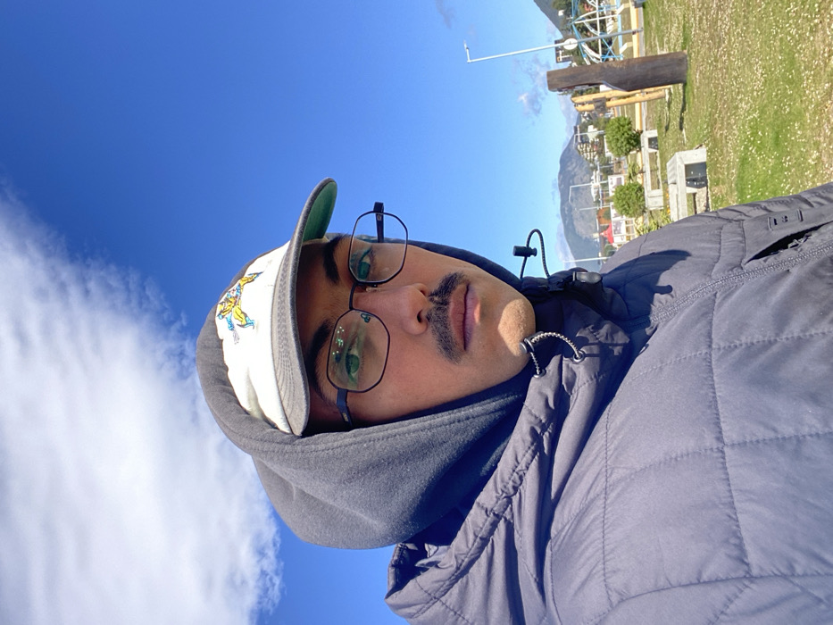 An adult male individual is depicted in a close-up, eye-level, selfie-style photograph, looking directly into the camera. He wears a dark, possibly charcoal, quilted puffer jacket over a dark grey hooded sweatshirt with the hood pulled up. A light-colored baseball cap featuring a yellow and blue graphic (resembling a comic book character) is worn under the hood. The individual has dark hair, a thin dark mustache, and is wearing oval-shaped dark-rimmed prescription glasses. Drawstring toggles from his jacket are visible below his chin.
The background reveals an outdoor public green space or park in Ushuaia, Argentina. The sky is clear blue with a singular, elongated white cloud formation positioned high on the left. The ground consists of short green grass dotted with small white flowers. In the middle ground, several light-colored concrete benches, rustic wooden playground structures, and a tall street light pole are visible. Further in the background, a street runs alongside various low-rise buildings. Beyond the urban structures, rolling, partially forested mountains rise under the bright daylight sky, characteristic of the region of Ushuaia.
