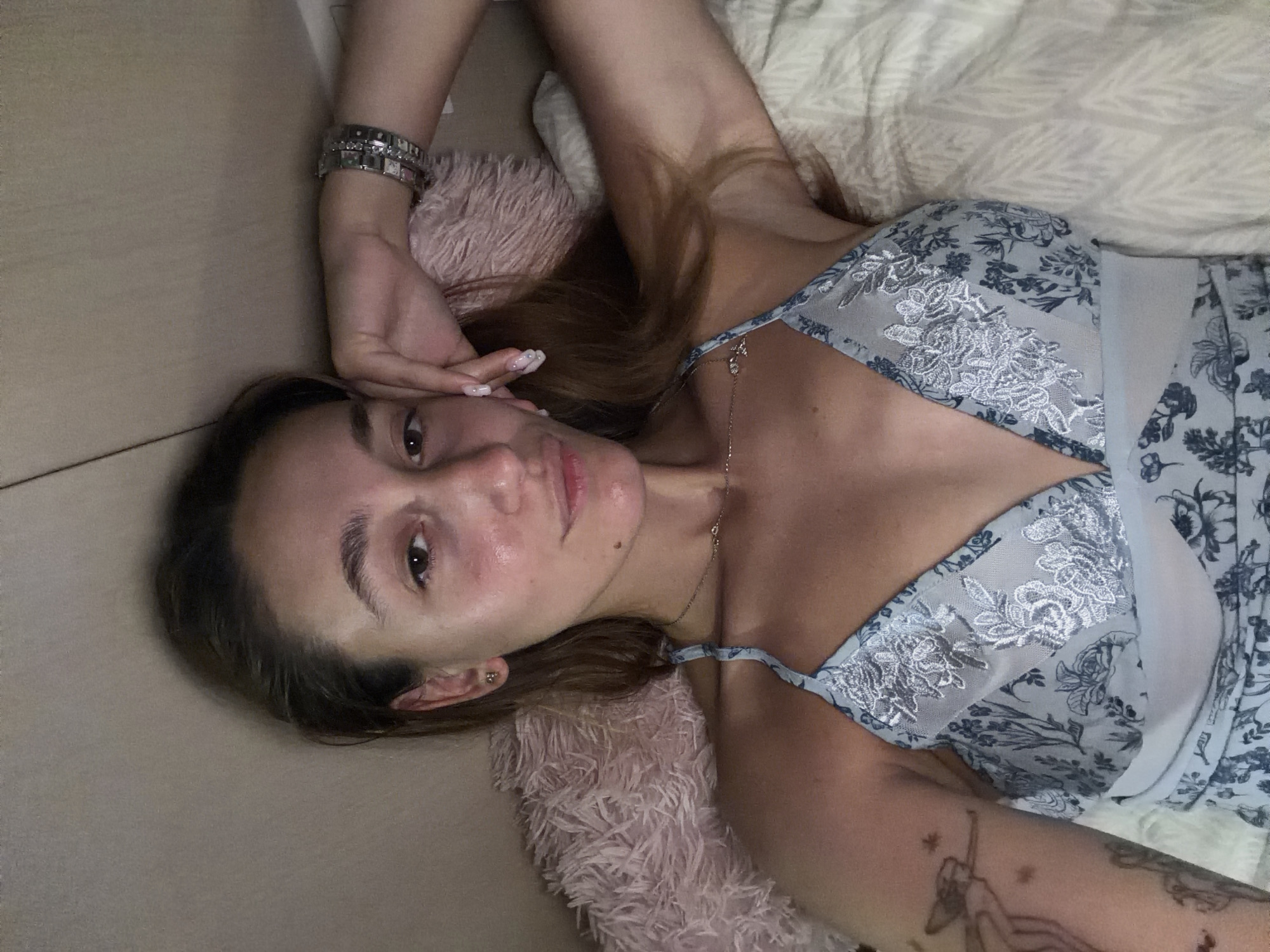 The image shows a woman lying on a bed, with her head resting on a pink, fluffy pillow. She is wearing a light blue slip with a floral pattern and lace detailing. Her right arm is bent, and her hand is resting on her cheek. A silver watch with multiple links is visible on her wrist. A delicate silver necklace with a small pendant is around her neck. She has dark hair, and her skin appears to have a slight sheen. There is a tattoo on her left arm. The background consists of a light-colored wall and bedding. The scene appears to be an indoor domestic setting, likely a bedroom. There are no explicit details to indicate the location is the city of Sofia, Bulgaria.