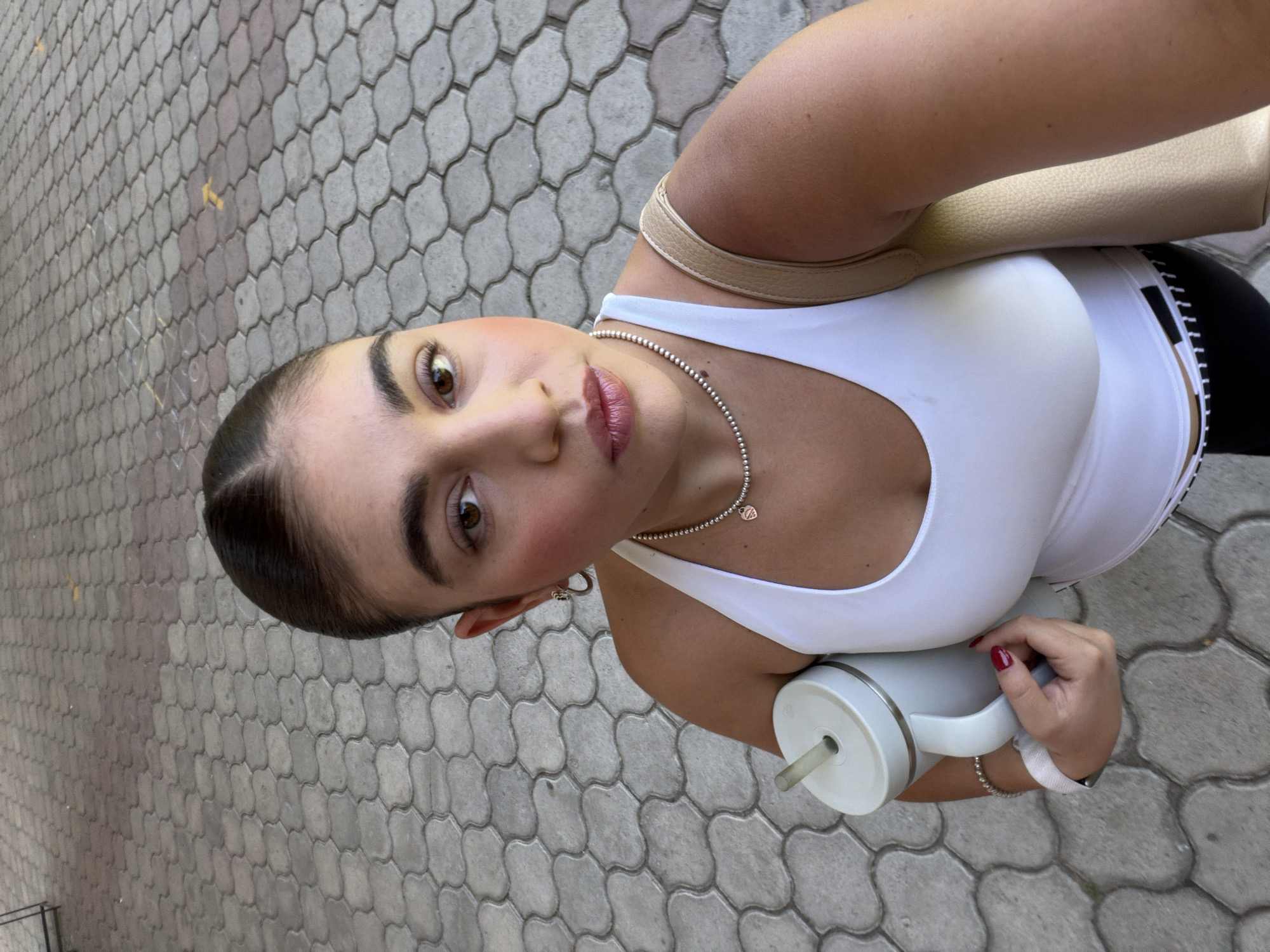 The image is a vertical, eye-level selfie of a person with feminine features, taken outdoors in San Bernardino Tlaxcalancingo, Mexico. The subject looks directly into the camera with wide, dark eyes and a neutral, slightly pouting expression, showcasing full eyebrows, blush, and pink lip gloss. Her dark hair is pulled back, revealing a clean hairline.
She is wearing a white athletic tank top, a silver beaded necklace with a small heart pendant, a silver bracelet, and a watch. A beige shoulder bag strap is visible over her right shoulder. In her right hand, which has bright red nail polish, she holds a light grey or white tumbler with a straw.
The background features distinctive grey interlocking paving stones, suggesting an outdoor, possibly urban or suburban, environment. The bright lighting indicates it is daytime. There is no visible text in the image.