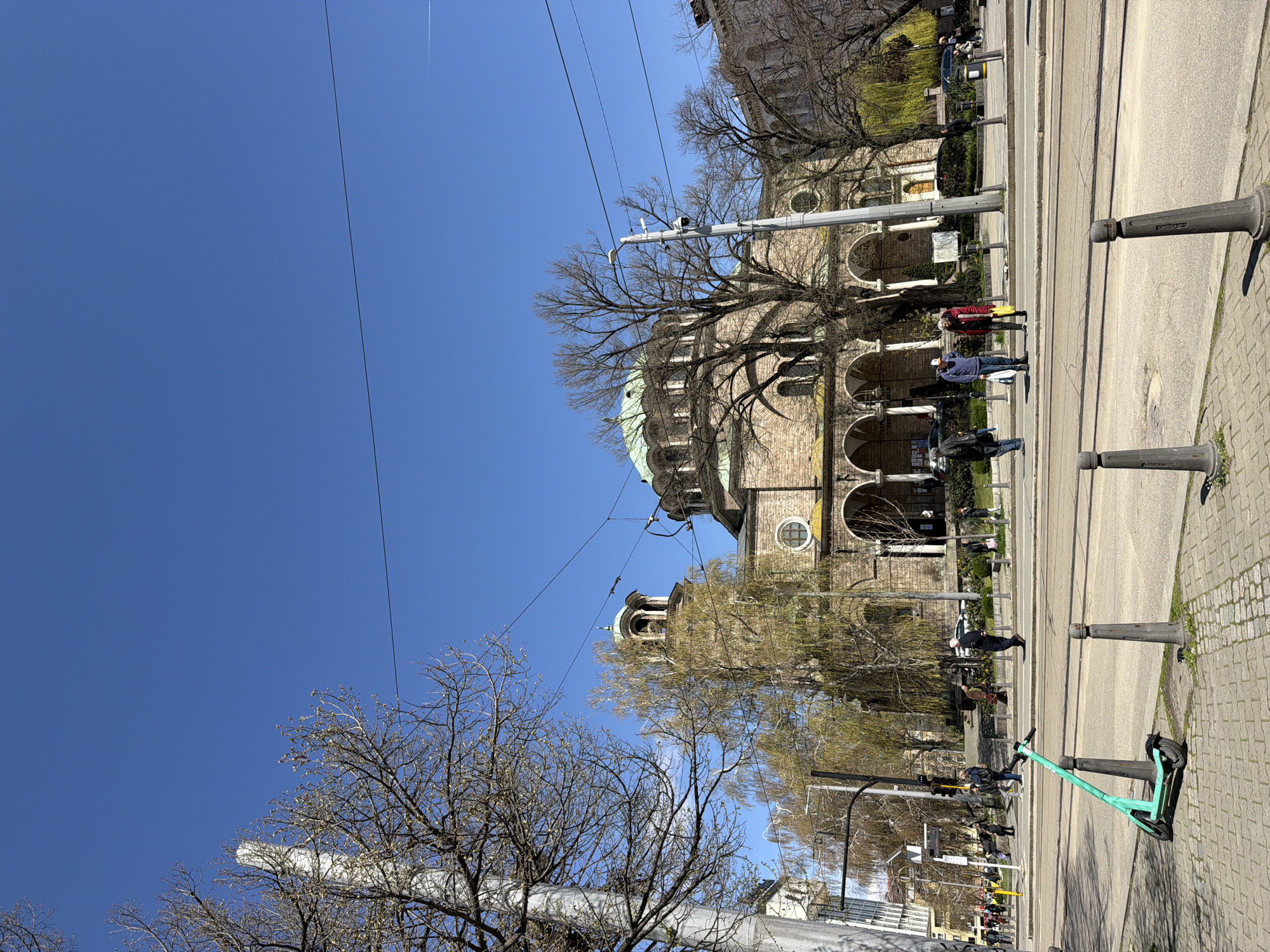 The image captures a street scene in Sofia, Bulgaria, featuring the Alexander Nevsky Cathedral. The cathedral, constructed of stone, has visible domes. The scene is shot on a sunny day under a clear blue sky, with power lines visible. The street is lined with trees. A parked green scooter is visible on the right side. People are walking near the cathedral's facade. Cast iron bollards are lining the edge of the road.