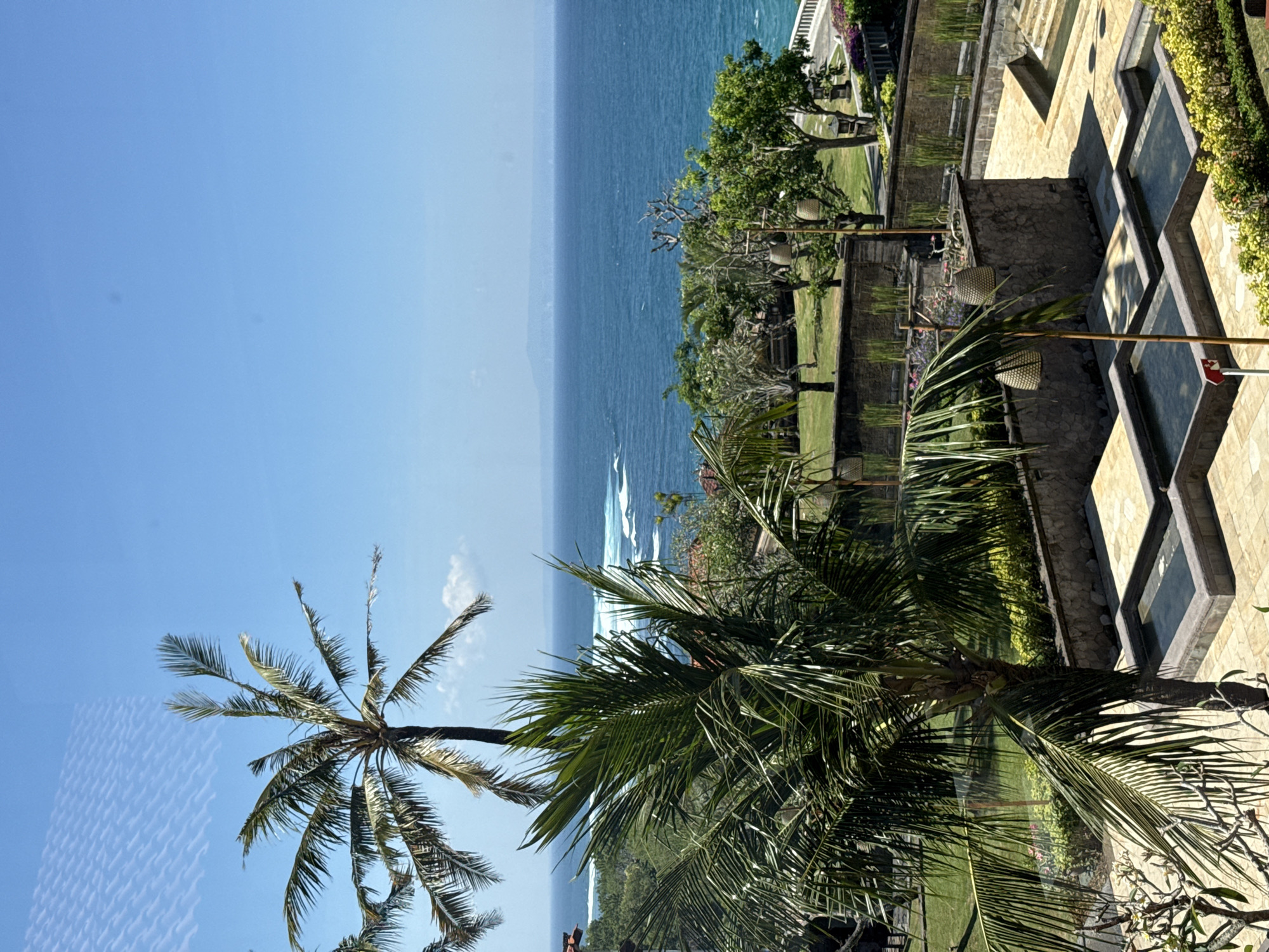 The image presents an elevated, sunlit view of a tropical coastal landscape in Kuta Selatan, Indonesia, from what appears to be a window or balcony, indicated by a faint grid-like reflection in the upper left sky. A clear, bright blue sky dominates the upper frame, meeting a distant, hazy blue horizon over a vast expanse of deep blue ocean. Far-off landmasses are faintly discernible on the horizon. Closer to the foreground, white-capped waves break on the ocean surface.
The midground is characterized by dense, verdant tropical foliage, including numerous palm trees with their distinct fronds, and various other green trees and shrubs. Manicured grassy lawns are visible between the vegetation and architectural elements.
The foreground features a multi-level, terraced garden and resort area constructed with textured stone walls and light-colored paved pathways. Several rectangular, shallow water features or pools are integrated into these terraces, reflecting the sunlight. Wicker-basket-style lamps are suspended from poles at various points within the gardens. A small red sign is visible on a pole near a foreground water feature. No individuals are present in the scene.