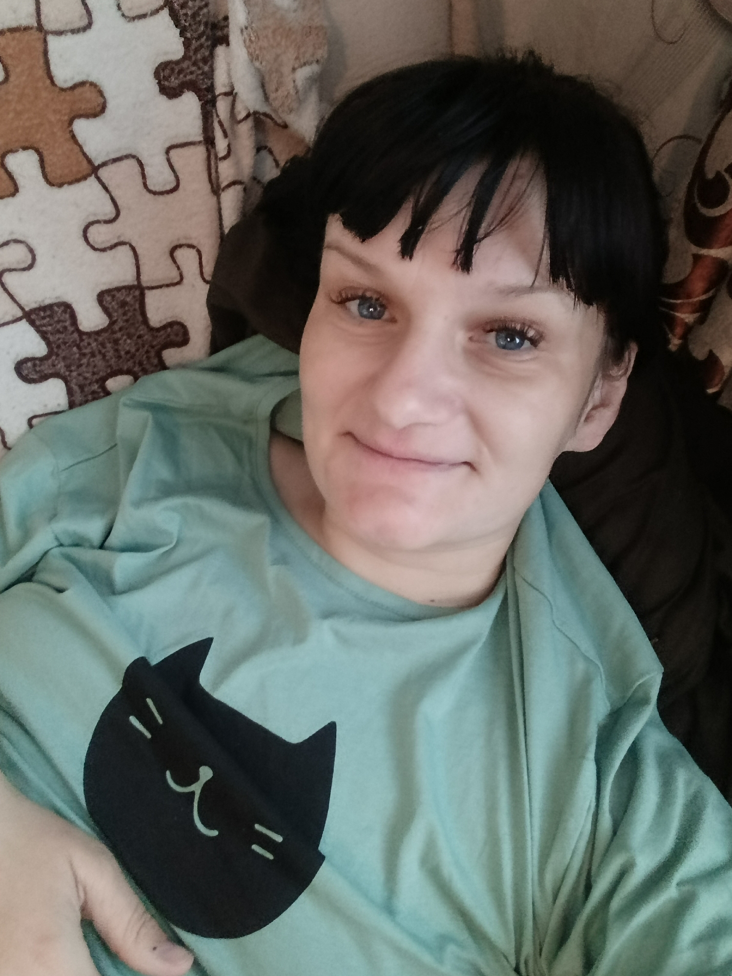 An adult woman is captured in a close-up, chest-up shot, looking directly at the camera with a gentle, soft smile. She has dark, short hair with bangs and striking blue eyes with long, defined eyelashes. Her complexion is fair.
She is wearing a light teal or mint green long-sleeved shirt. Prominently displayed on the front of her shirt is a black, stylized cat head patch. The cat patch features minimalist white details for its face: two small horizontal lines for eyes, an upside-down 'v' shape for a nose, a curved line for a mouth, and two short horizontal lines on each side suggesting whiskers.
The woman appears to be leaning back against something, possibly a cushion or headboard. The background features a patterned fabric with an interlocking jigsaw puzzle piece design in shades of light and dark brown, and off-white. To her right, a darker, possibly solid brown or black fabric is visible, suggesting a couch, bed, or another piece of furniture.
The overall scene suggests a relaxed, indoor setting, likely a home in Фоки, Russia. The lighting is soft and even, indicating an absence of harsh direct light, though the exact time of day is not discernible. The woman's expression conveys a sense of contentment or ease. No visible text is present in the image.