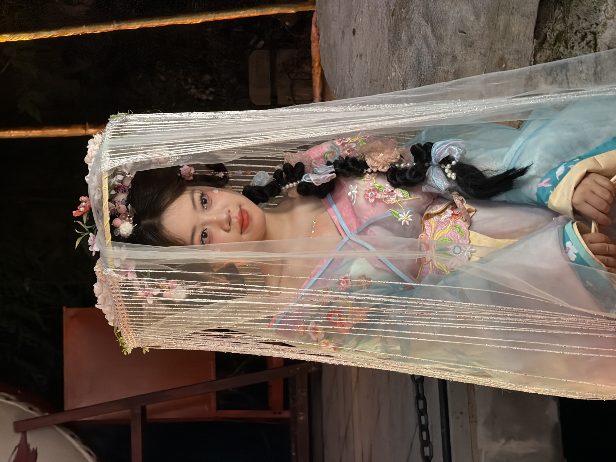 A young East Asian woman with dark hair styled in braids and adorned with flowers and pearls, is captured in a medium shot. She is wearing a pale pink and blue traditional Chinese garment, possibly a Hanfu, which is embellished with floral embroidery. She is partially obscured by a sheer, white veil or canopy decorated with small flowers and beaded strands, which frames her head and upper body. Her makeup features pink blush and a soft orange-red lipstick. She is looking slightly to her right, with a gentle expression.
The background is dark and appears to be outdoors at night. To the left, a reddish-brown wooden structure with diagonal supports is visible, suggesting some form of stage or seating. To the right, two vertical elements, possibly illuminated pillars or tree trunks, glow with a warm orange light. The ground beneath her appears to be a rough, textured surface, possibly stone or concrete. The overall scene suggests a performance or a themed event, possibly in an outdoor setting within Yuzhong District, China, known for its historical sites and cultural activities.